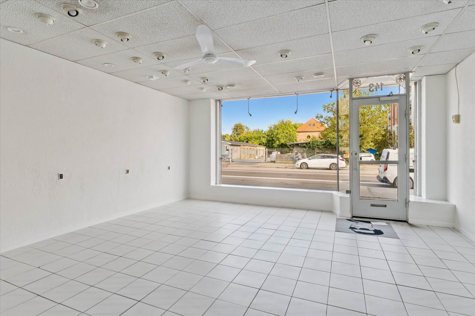 Lokale med hvidt loft, væg og gulv samt store vinduer med skygger på gulvet og glasdør med refleksioner fra biler udenfor