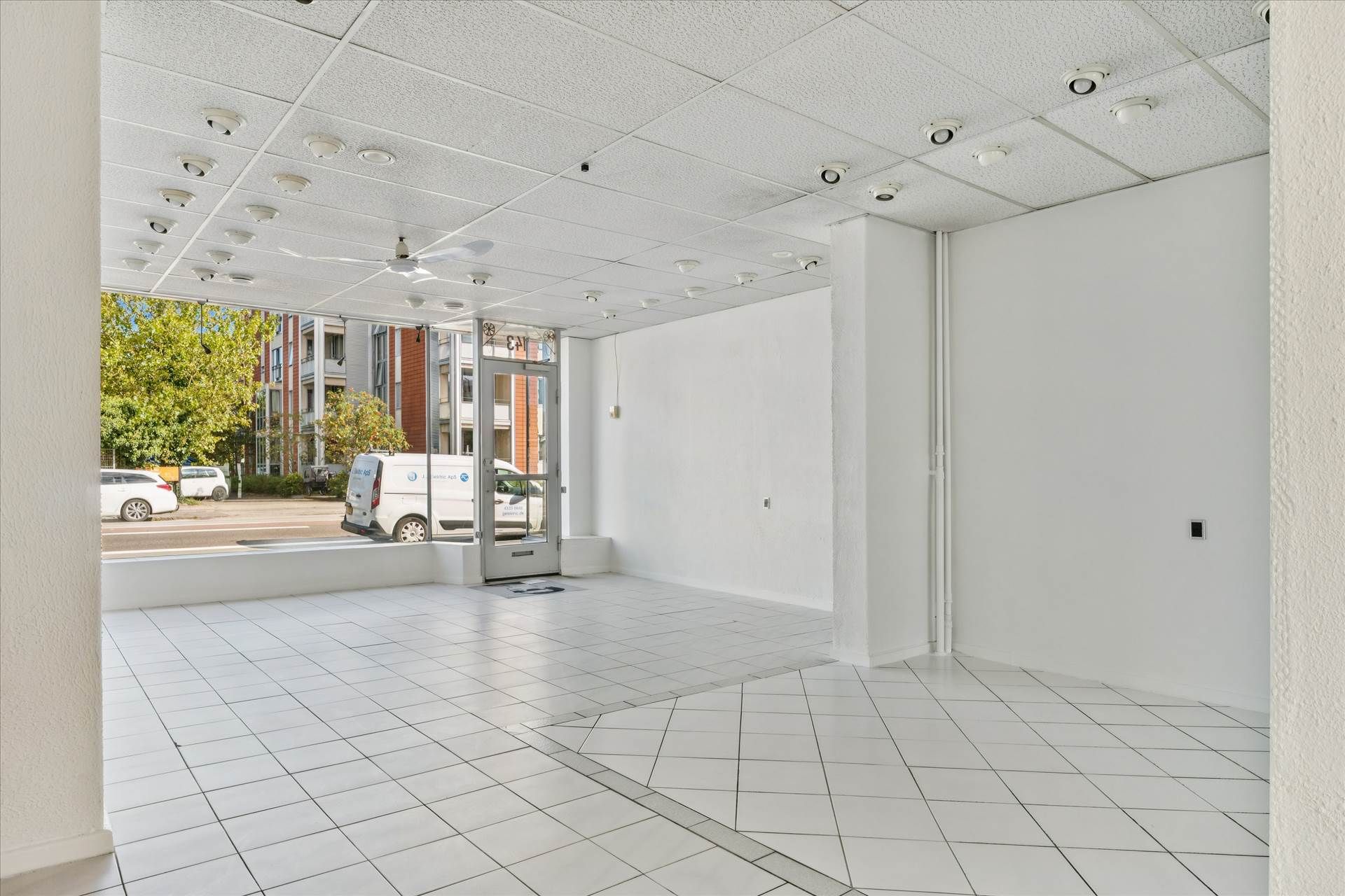 Lokale med hvidt loft, hvide vægge og hvidt gulv. Adgang til gaden med store ruder. Parkerede biler udenfor.