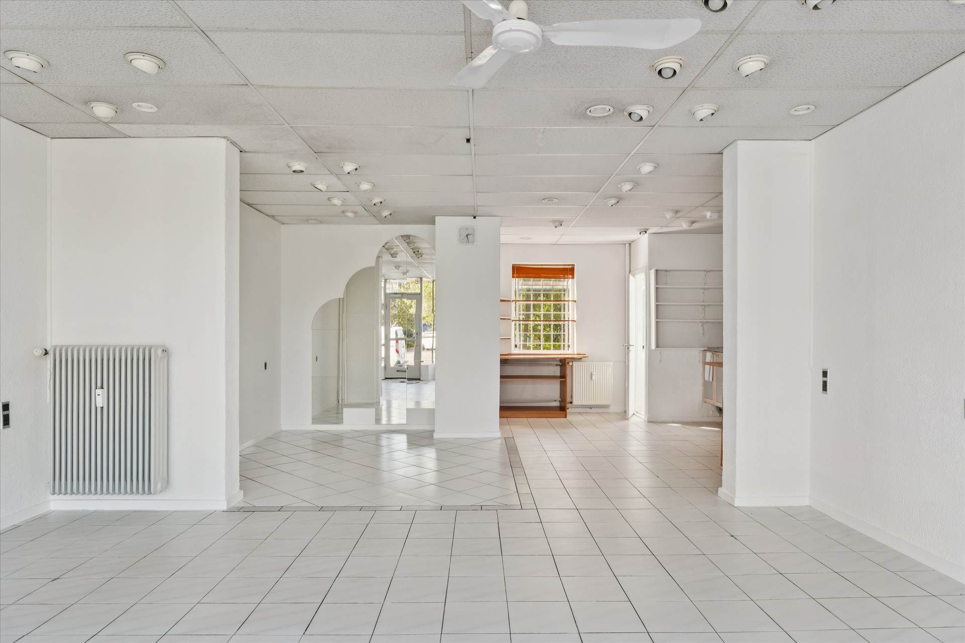 Foto af et lyse og rummeligt lokale med hvide vægge og gulvtæppe, gardiner og radiator, lyst trægulv og hvide lofter med