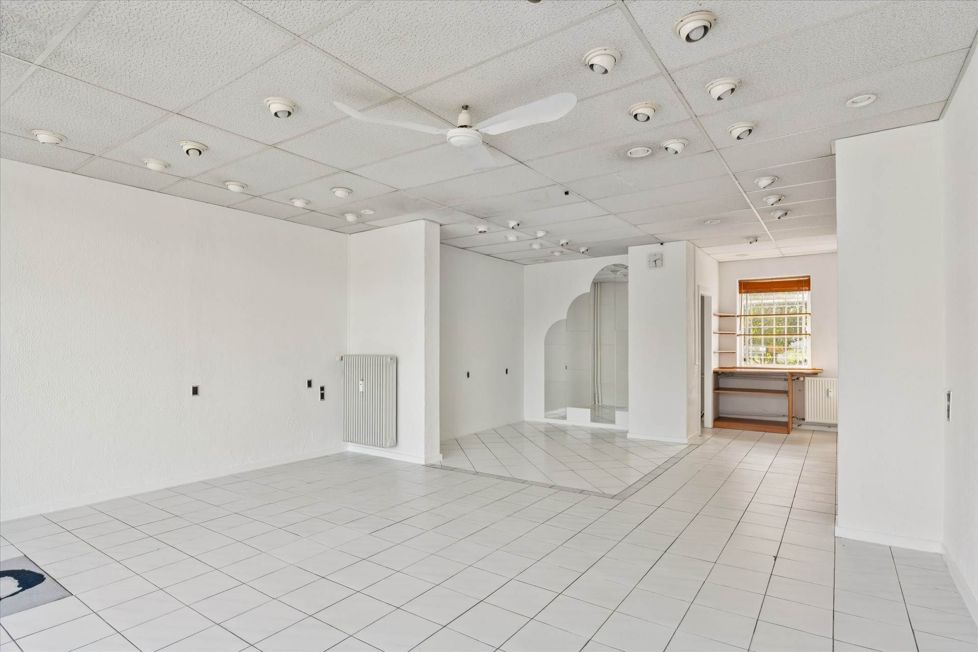 En lys og rummelig lokale med hvide vægge og gulvfliser, et hvidt loft med ventiler og en ventilfan. Et hvidt hjørnekabinet