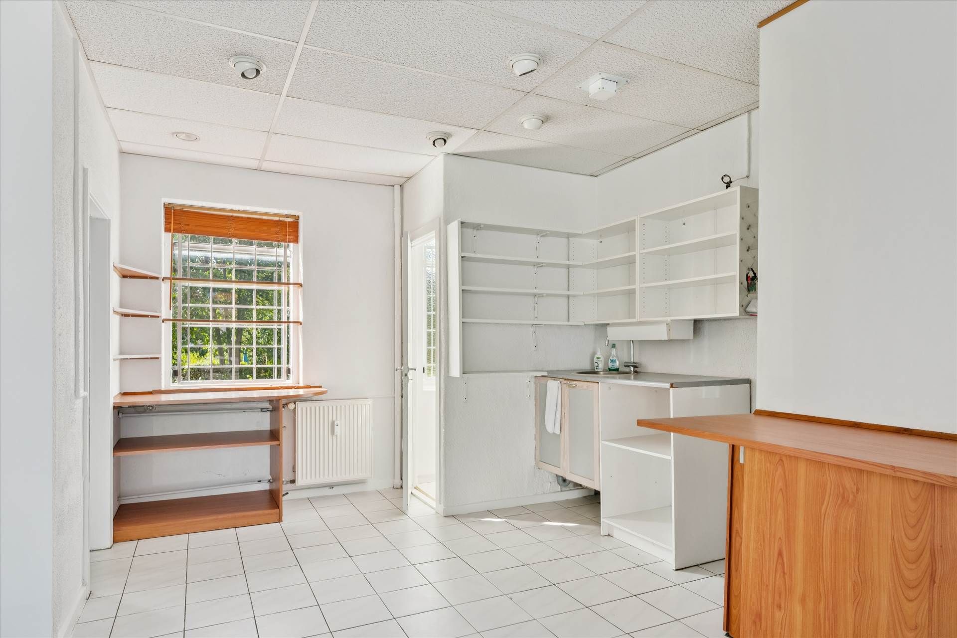 Lokale med hvide vægge og gulv, åbent køkken med højre dør, hældende gulv, lyse vinduer, indbygget køleskab og skraldespand