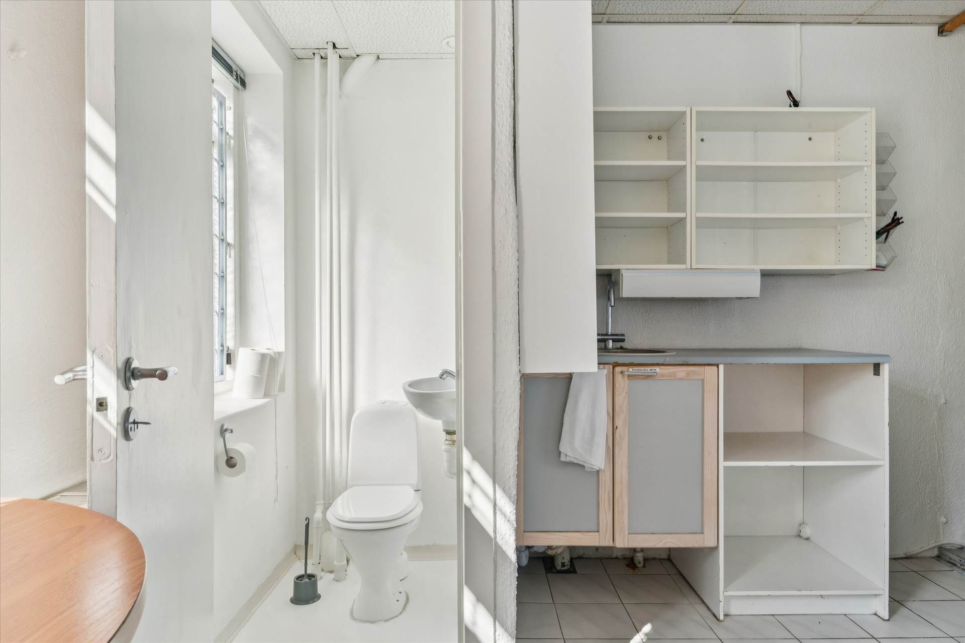 Et lille badeværelse med hvide vægge, toilet, håndklæde, to toiletpapirruller, og et lille køkken med hvide hylder,