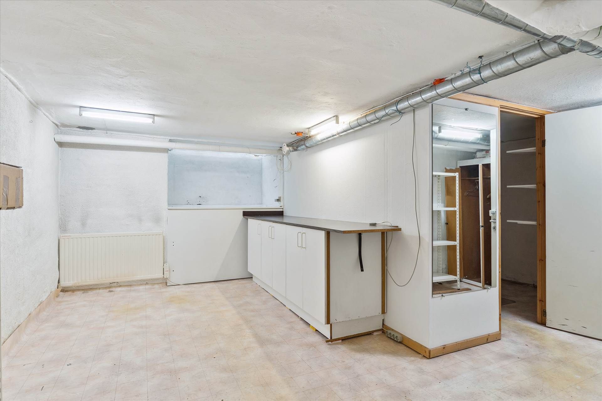 Rummeligt lokale med vinduesparti og radiator i hvidt værelse med højt loft og lyse gulvfliser samt åbne døre til skabe