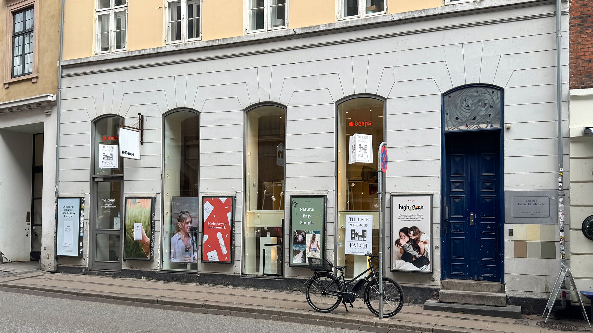 en cykel parkeret uden for en butik med navnet Denps. Butikken har flere vinduer og reklameskilte med tekst og billeder. Der