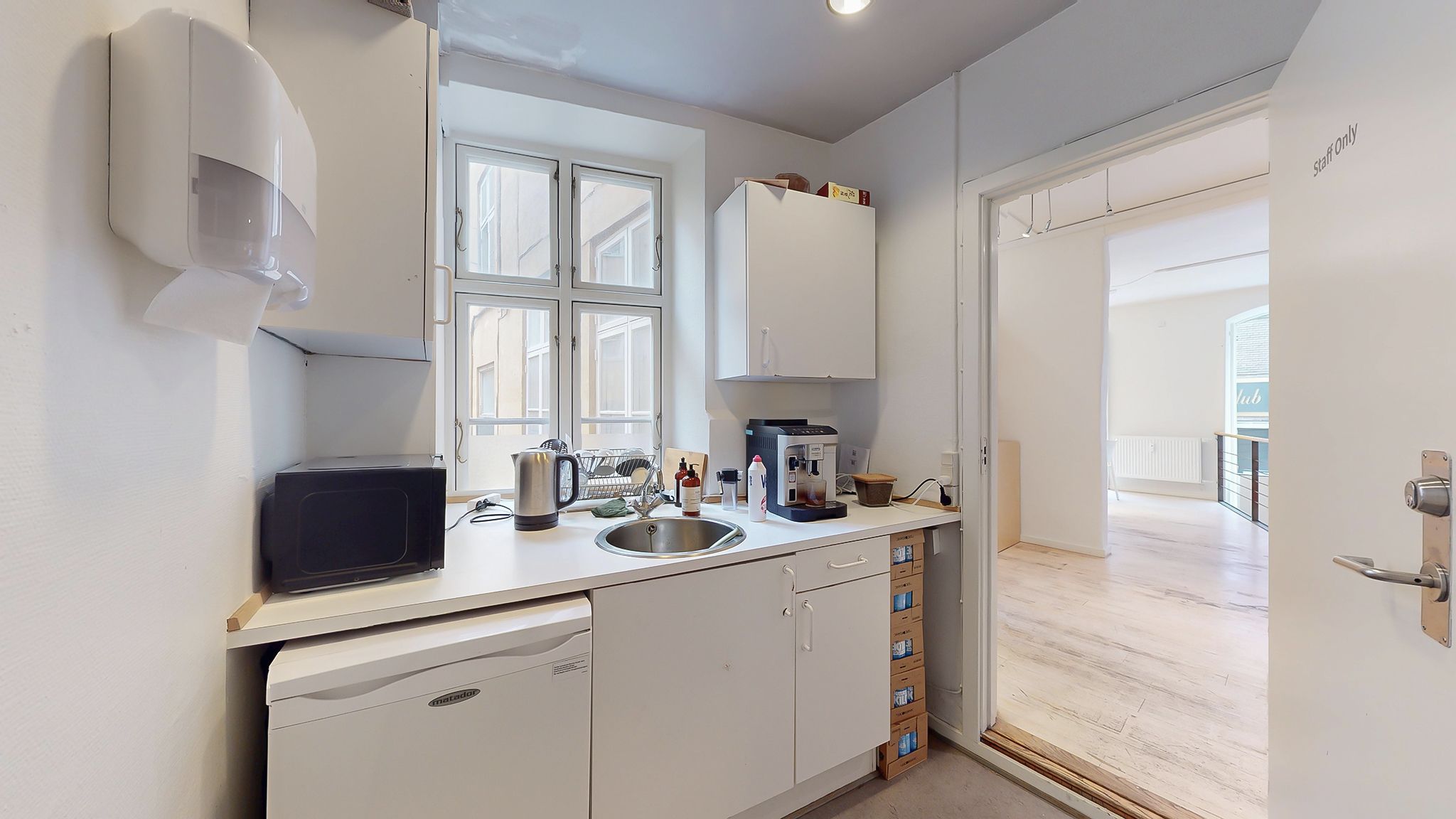 Køkken med hvide vægge og loft og moderne møbler samt apparater som en kaffemaskine, en mikrobølgeovn og en vaskemaskine