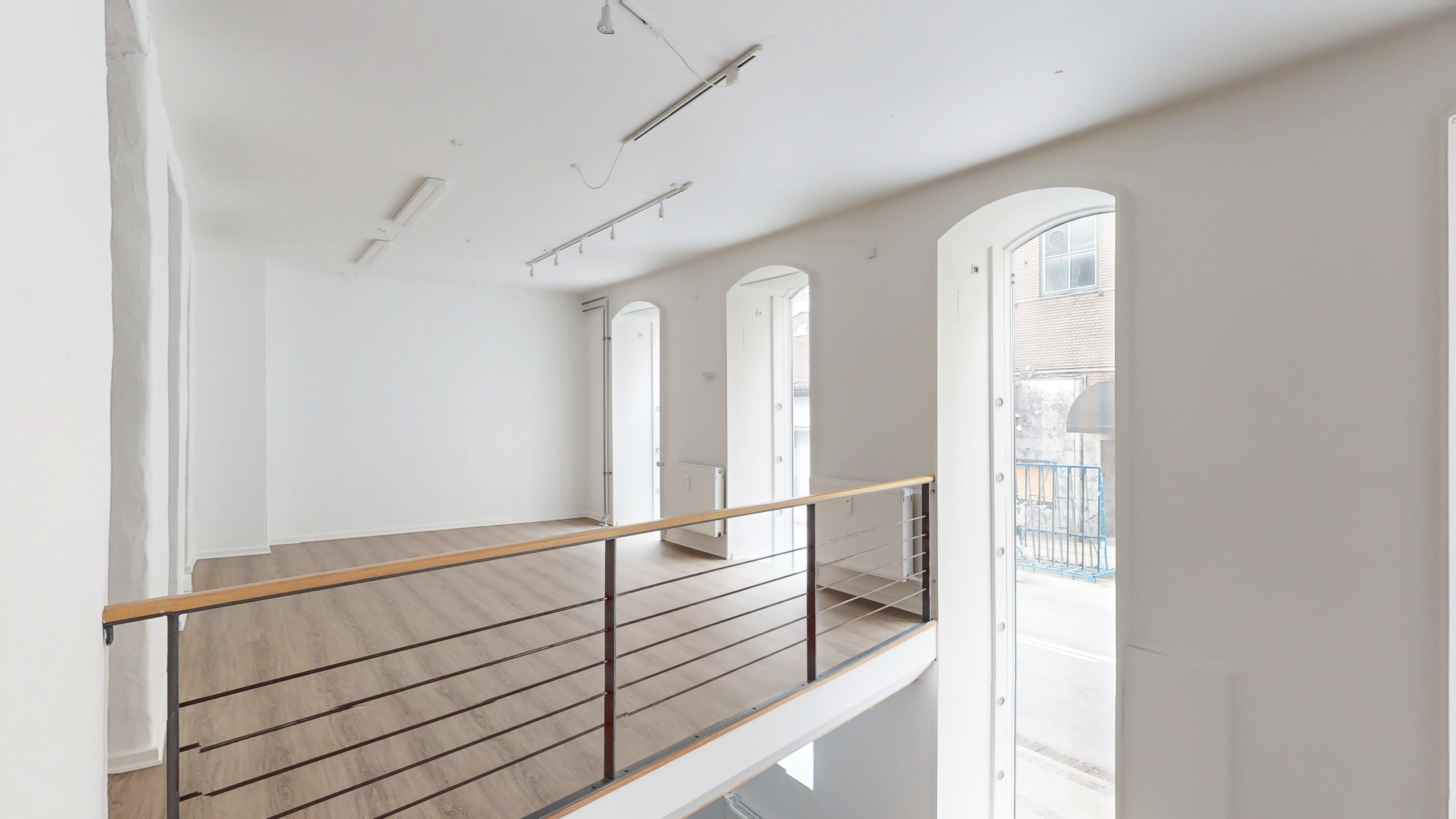 Store rum med højloft og parketgulv. Glasdøre og høj vinduesdekoration. Loftsmonterede spotlys og hvide vægge. Balkon med slanke stålstiger.