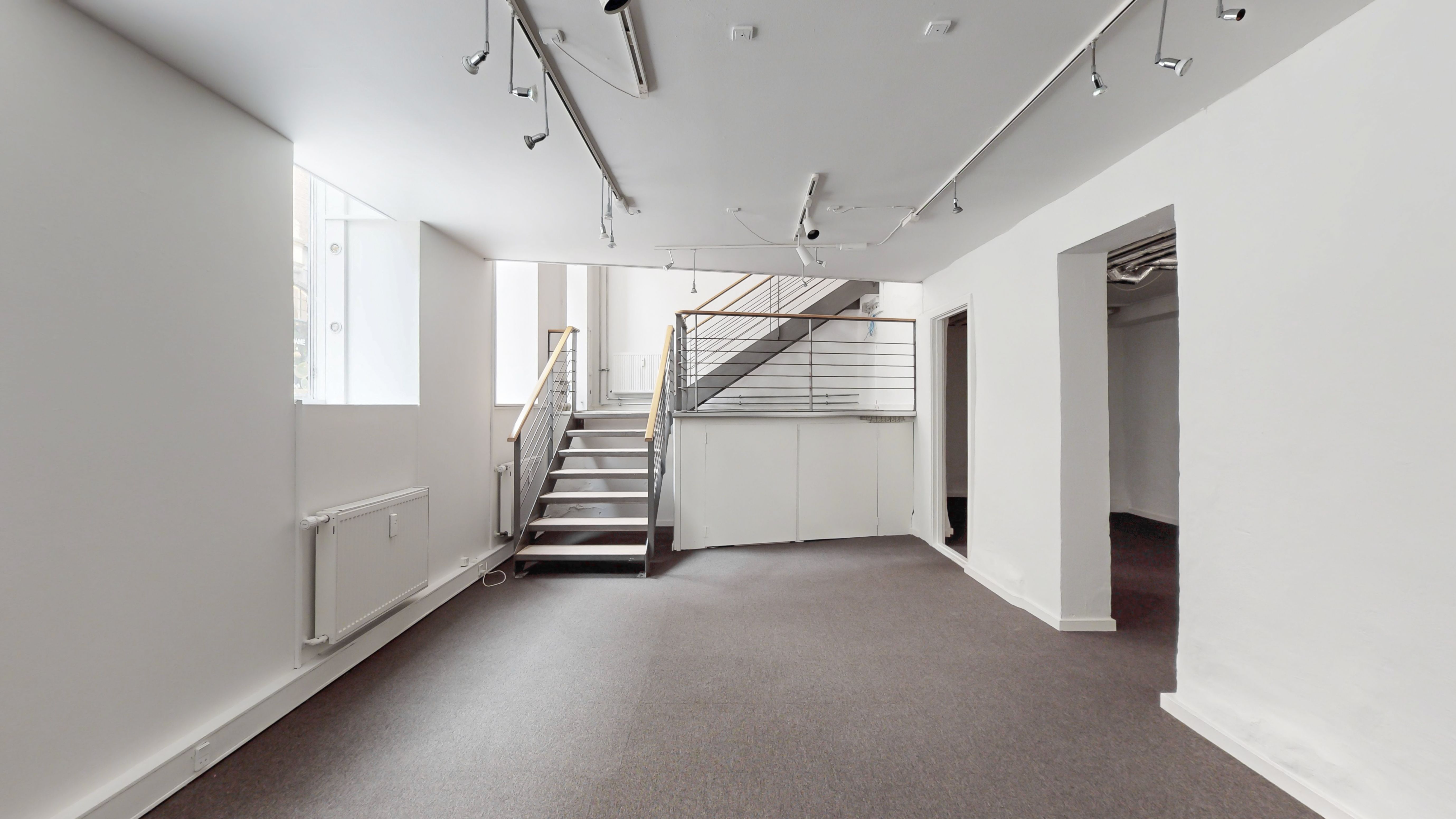 En stor og lys stue med lysegulve, lyse hvide vægge og lysekroner. Stigen med metalgevær og spidse trin er placeret til højre. Der er et vindue til venstre.