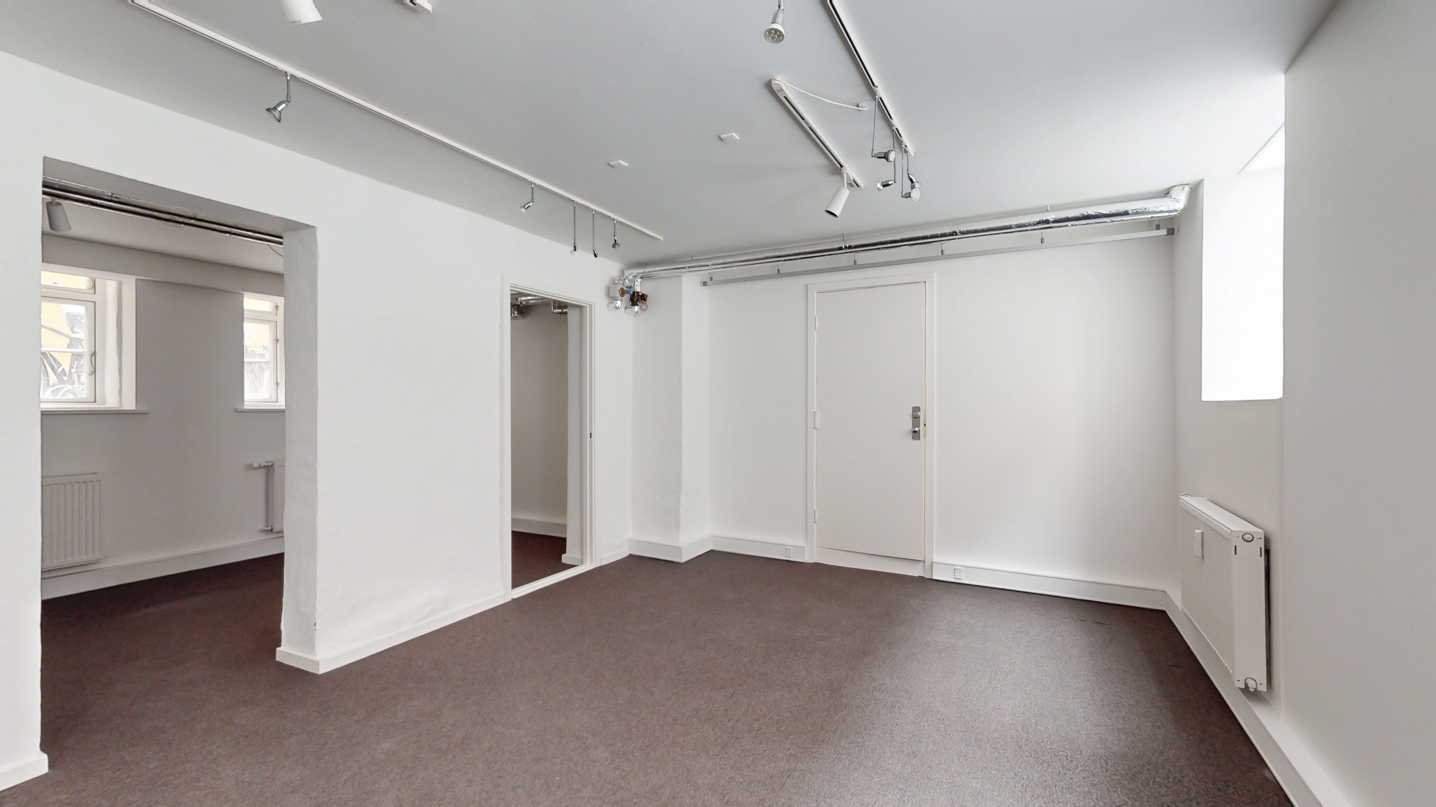 En stor og lys lagerlokale med hvide vægge og gulv. Der er to døre, der fører til et lille lagerrum. Loftet er udstyret med hængende spotlys.