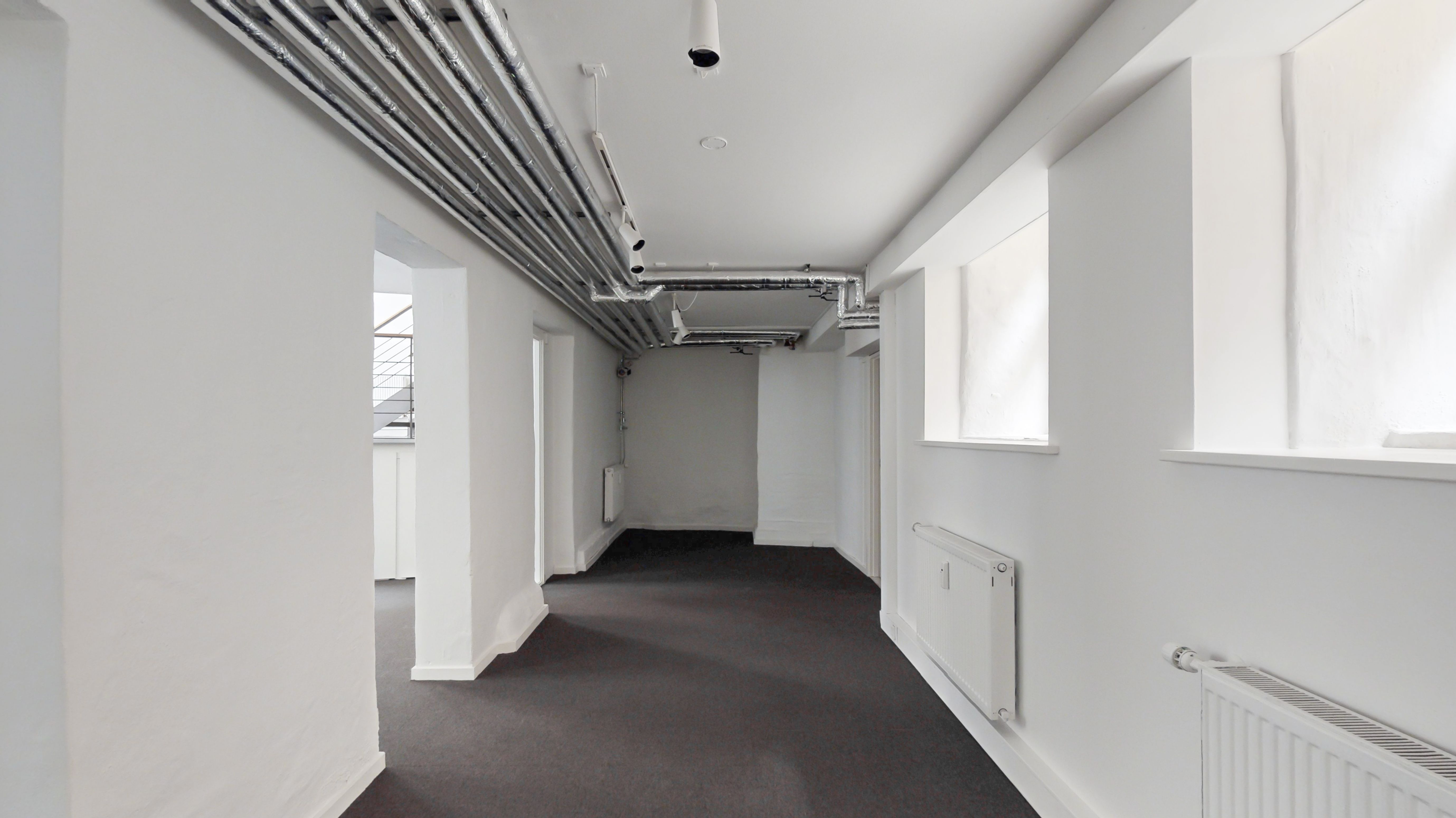 En ren og nyrenoveret korridor med hvide vægge og gulv og sort gulvbelægning og store vinduer og røre og piper på loftet