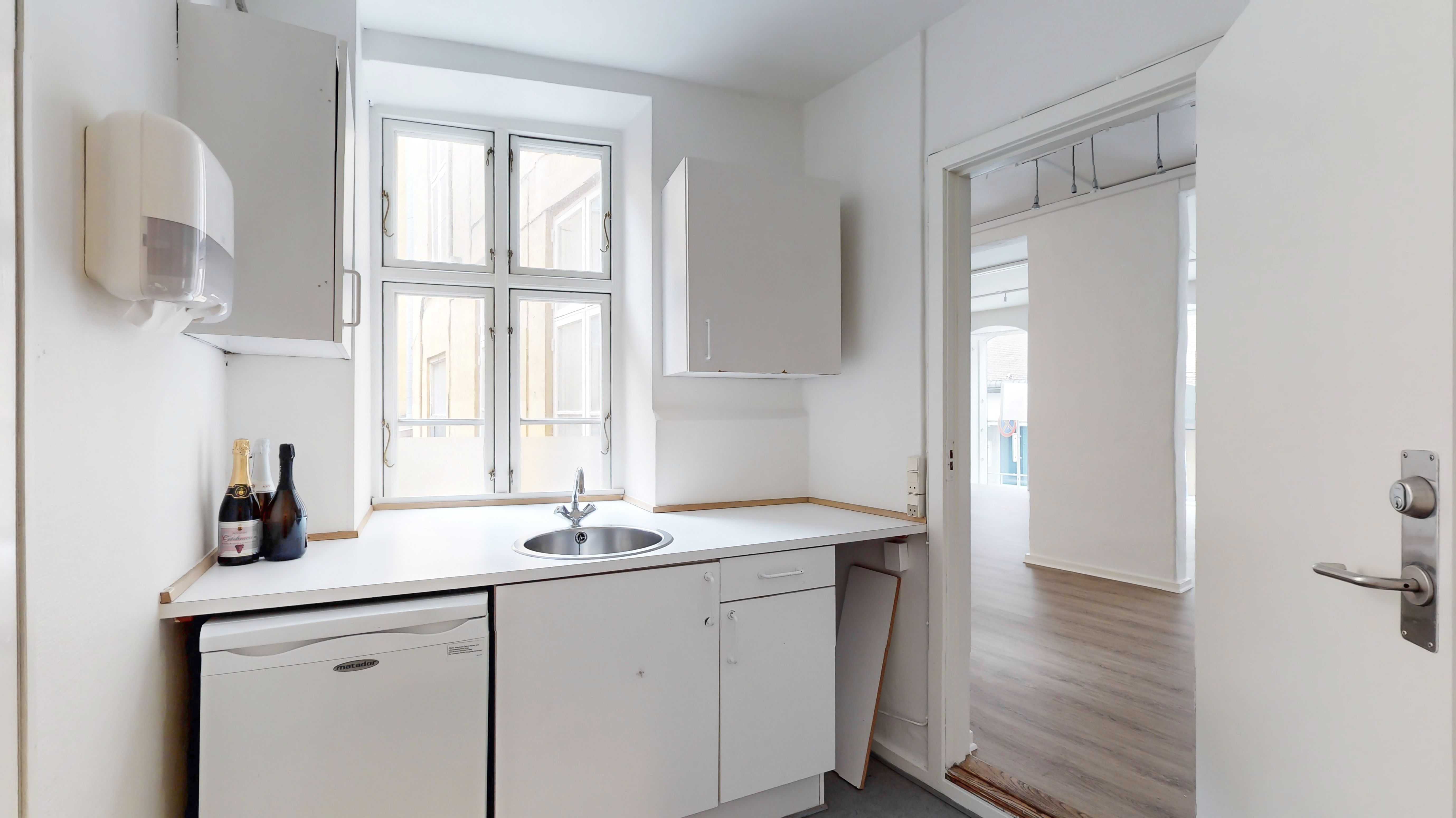 En lille og moderne køkken med hvide vægge og gulv, en håndvask med vandhaner og to champagneflasker på skraldespanden, samt en vaskemaskine og et køleskab
