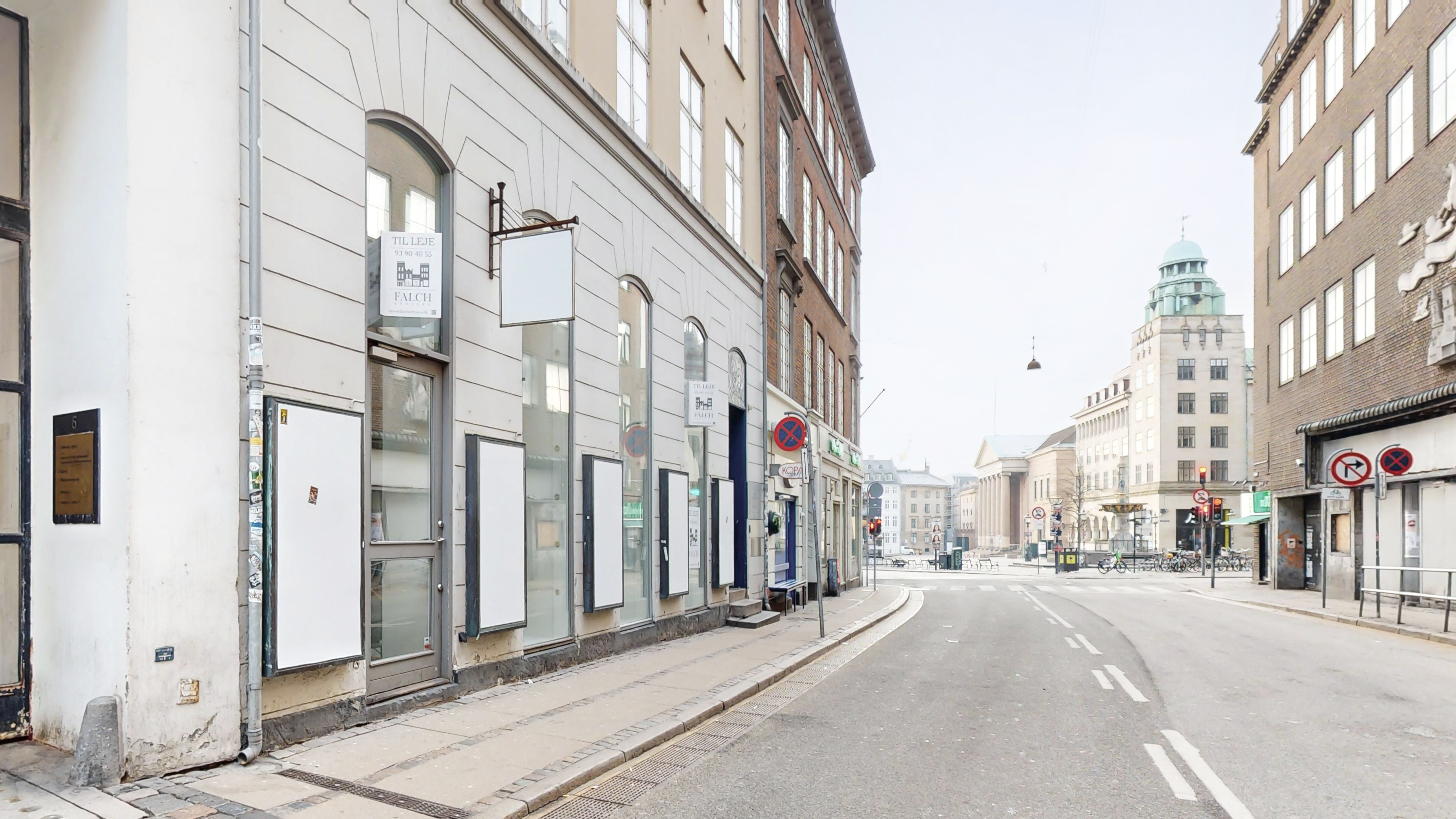 En tom gade med forretninger og skilte på hjørner i København. De fleste forretninger er lukket, og der er få mennesker på gaden.