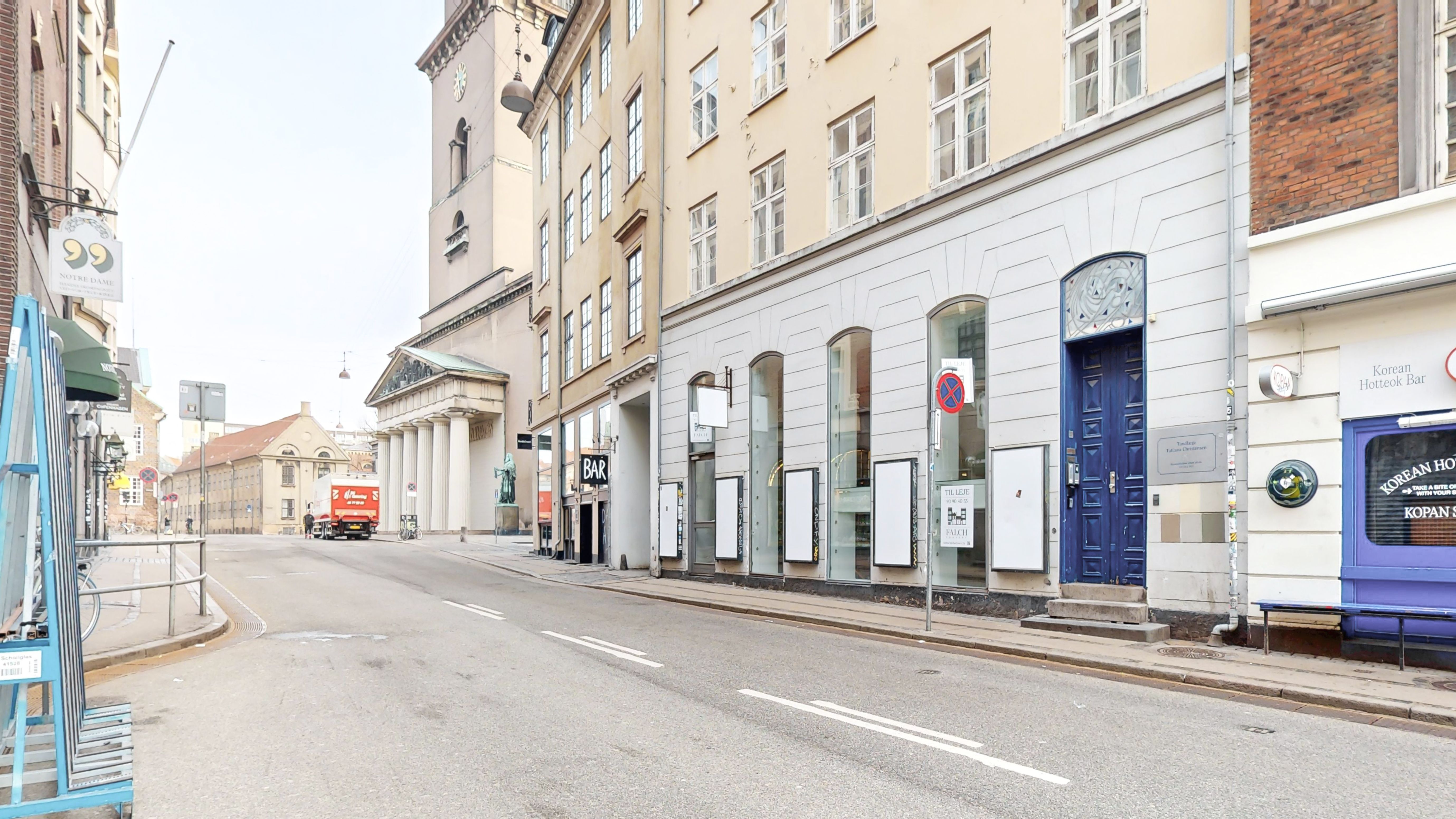 En bred gade med en kørende rødgrøn fragtvogn, hvor der er en koreansk hottoek bar og en detaljhandel med et blåt dørblad og et hvidt skilt med teksten Bar