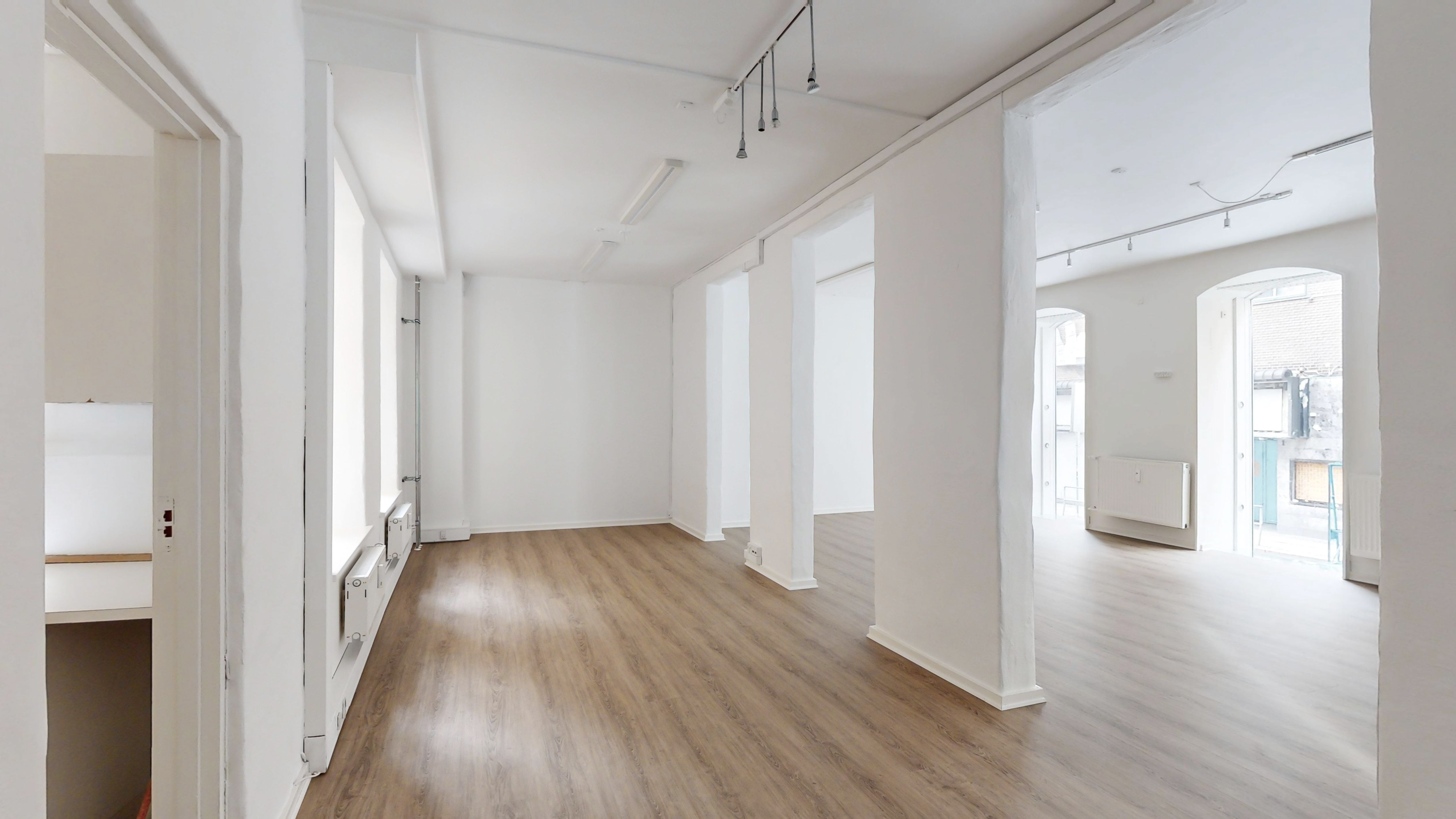 Det store lokale i hjertet af Indre By har lyse gulve og hvide vægge. På loftet hænger flere lysekroner og en lampe. Der er døre på hver side og et vindue i baggrunden