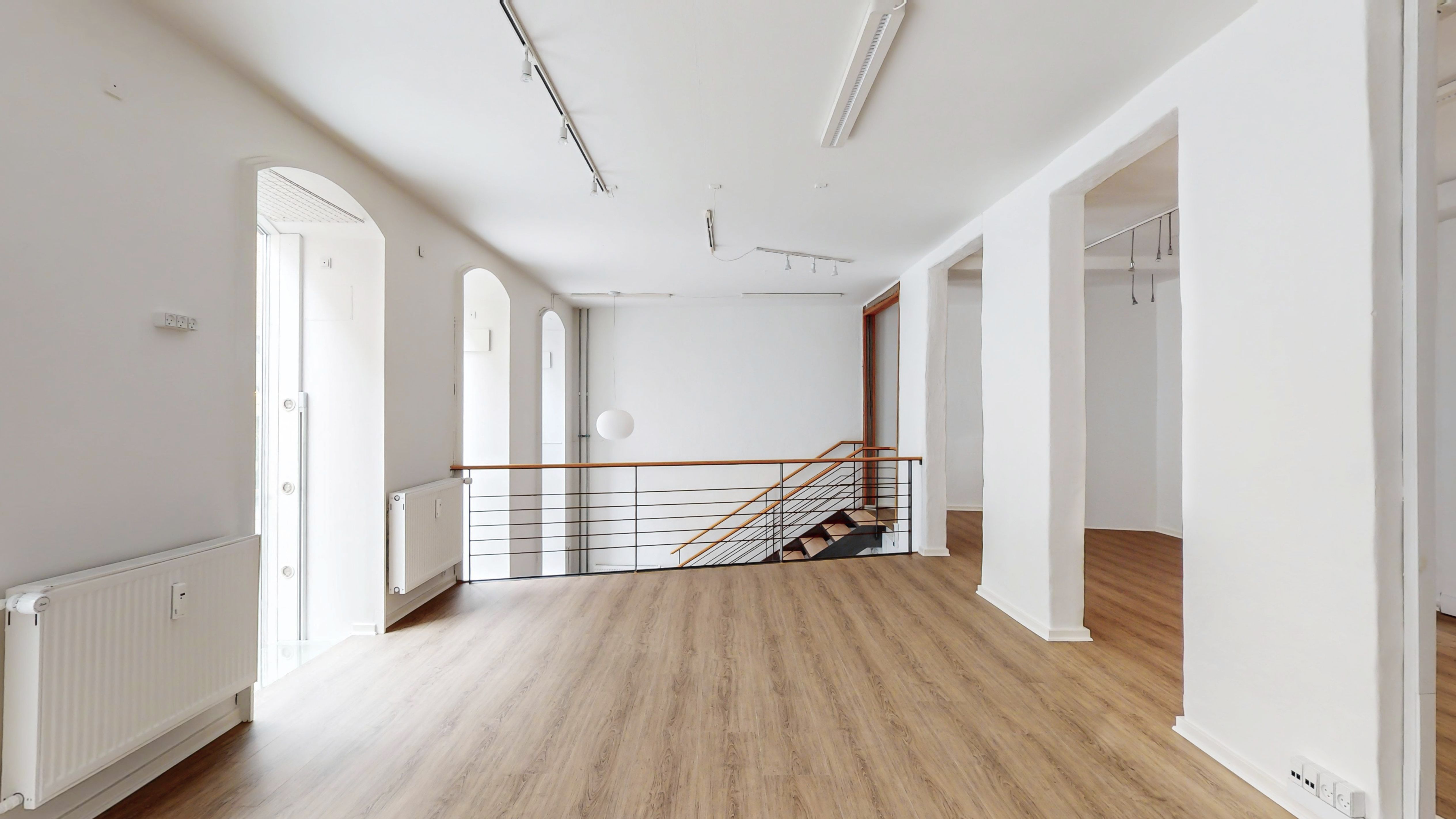 Hvidmalet rum med moderne højloft, lysekroner, lyse hårde trægulve og kælder. På højre side er der en moderne trappe med lyse stige og håndtag