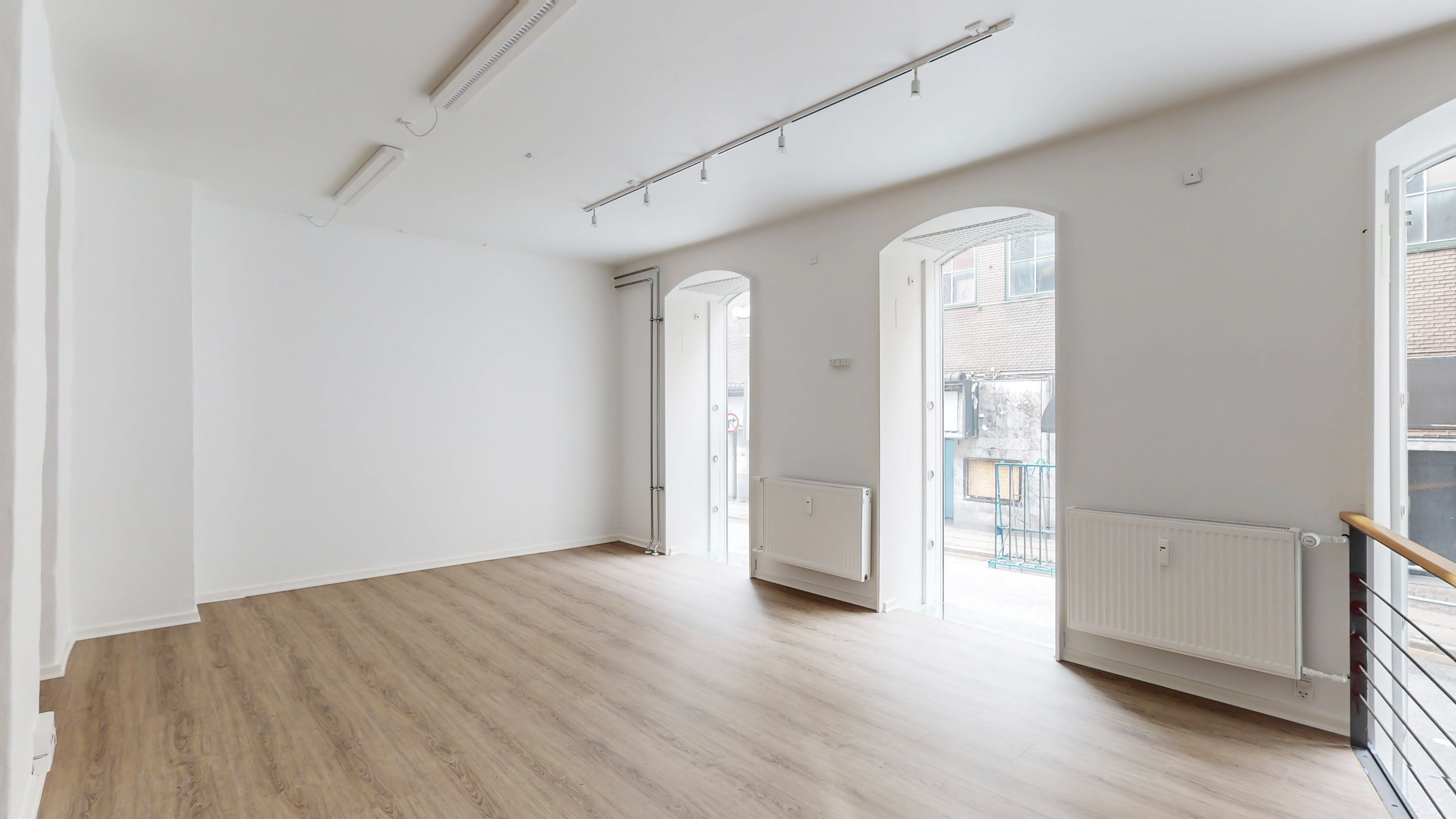 Et stort, lyse og tomt lokale med hvide vægge og lysegrå gulvbelægning. To vinduer og døre med bueformede overhoveder. To radiatorer og en åben balkon med metalgevær.
