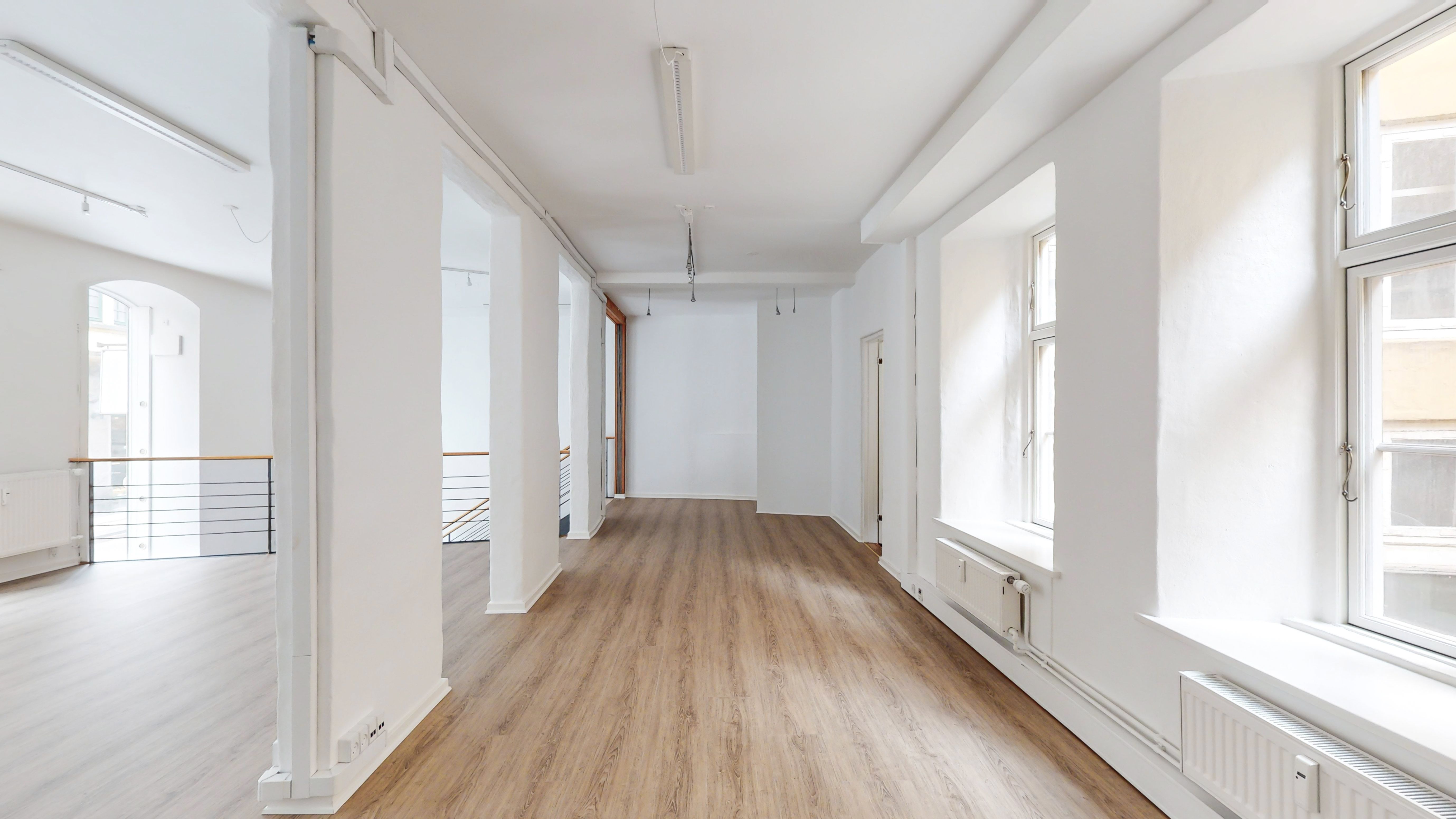En lang, ren og hvidt malet korridor med lyse trægulve og hvide vægge. Vinduer med hvide indramninger og et hvidt radiator. Lamper hængende på loftet og håndtag på en trappe.