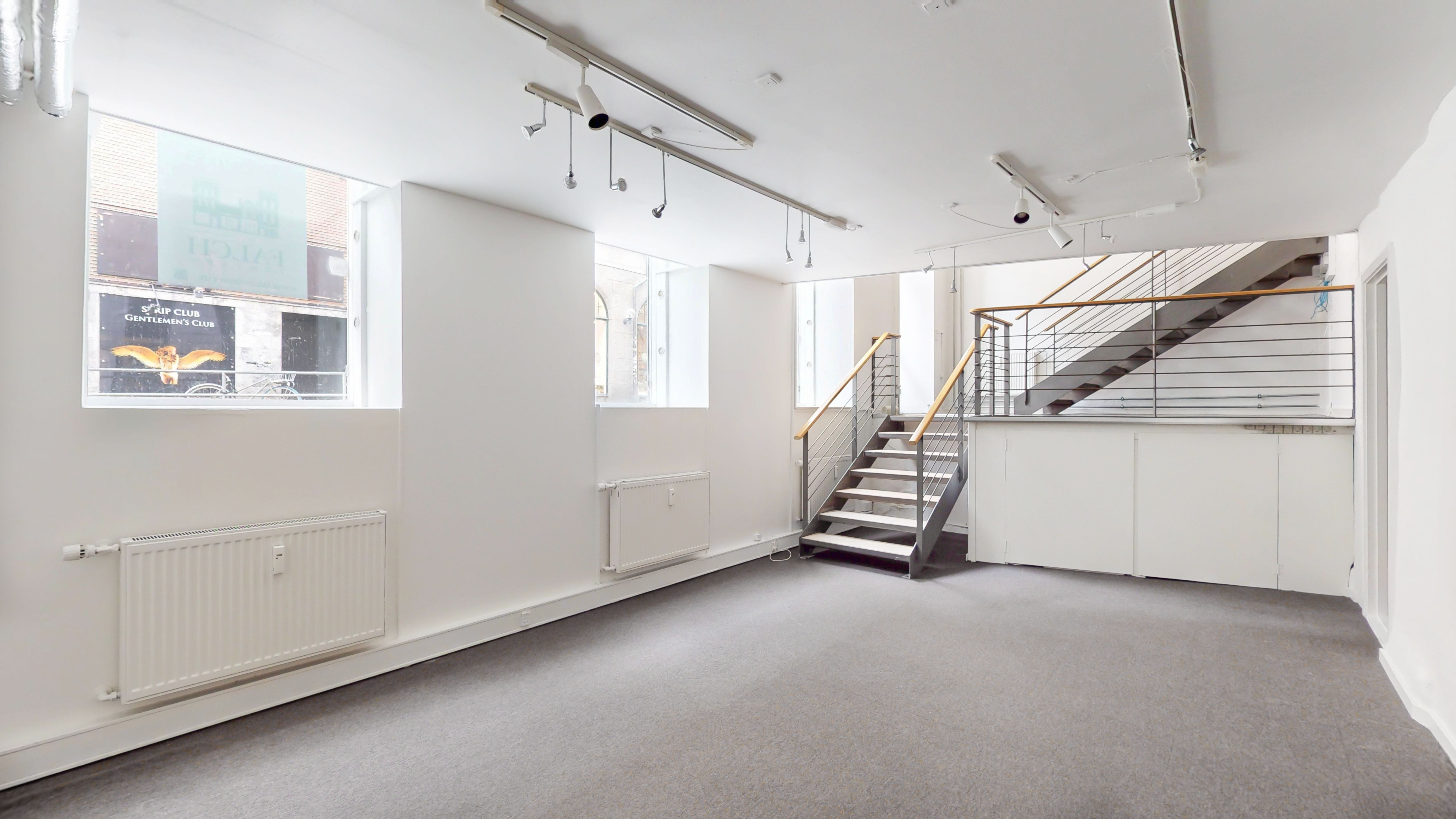 Store lyse vinduede lokaler med lysegul gulvbelægning og hvide vægge, to radiatorer og et trappe op til øverste etage med metalgeværer og trætrin