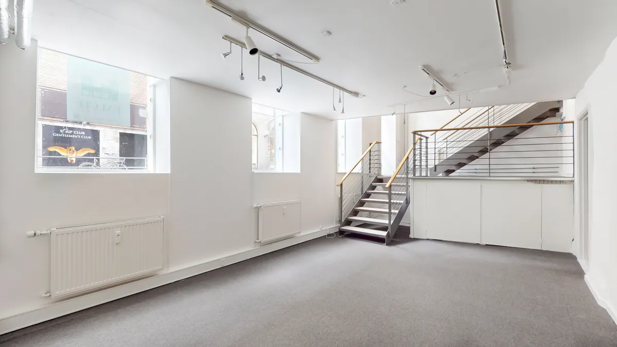 Store lyse vinduede lokaler med lysegul gulvbelægning og hvide vægge, to radiatorer og et trappe op til øverste etage med metalgeværer og trætrin