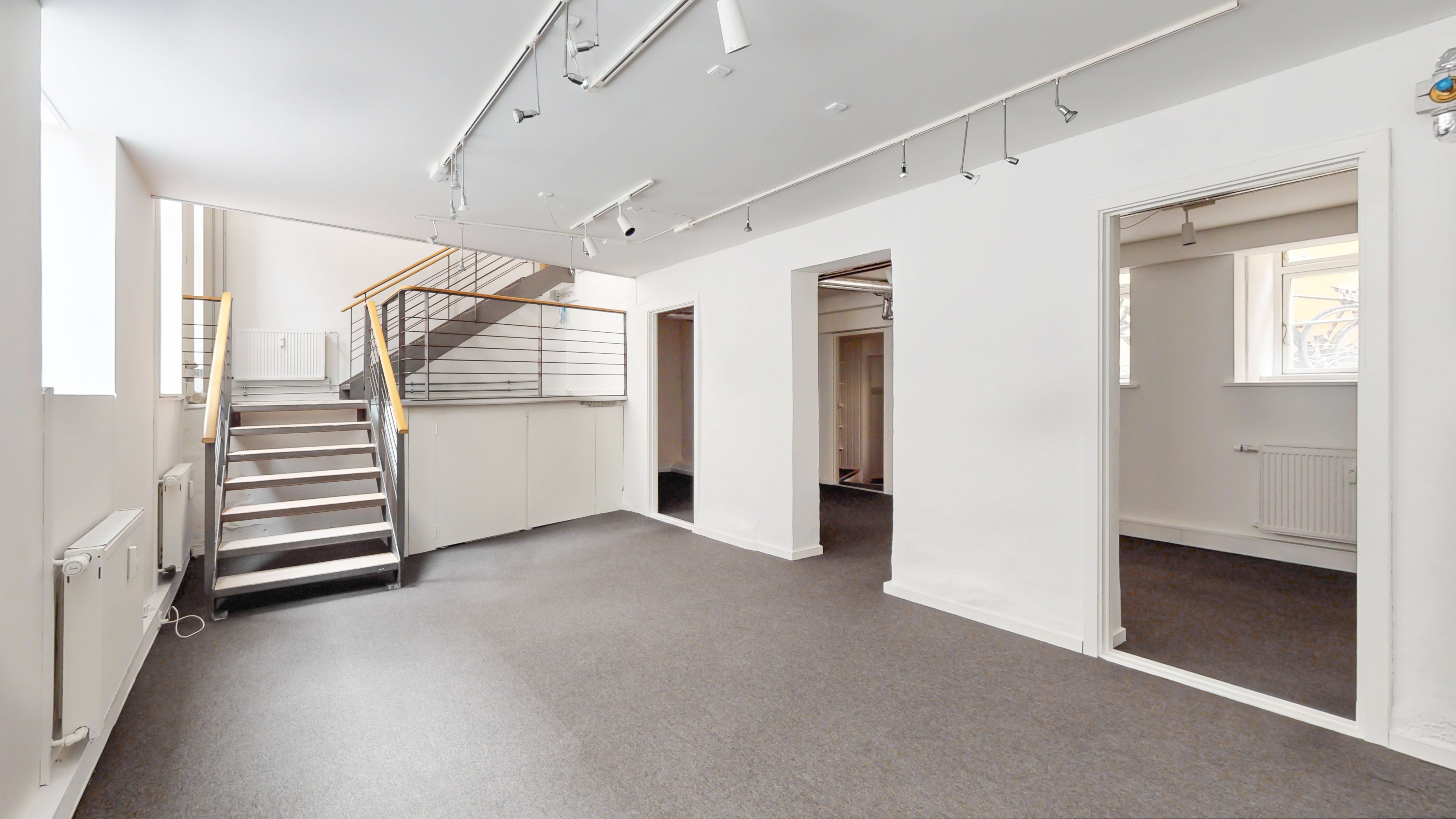 Et tomt lokale i stueplan med lyse loftsspots og lysegrå gulvbelægning. Der er en stige i hjørnet og et åbent vindue til højre.