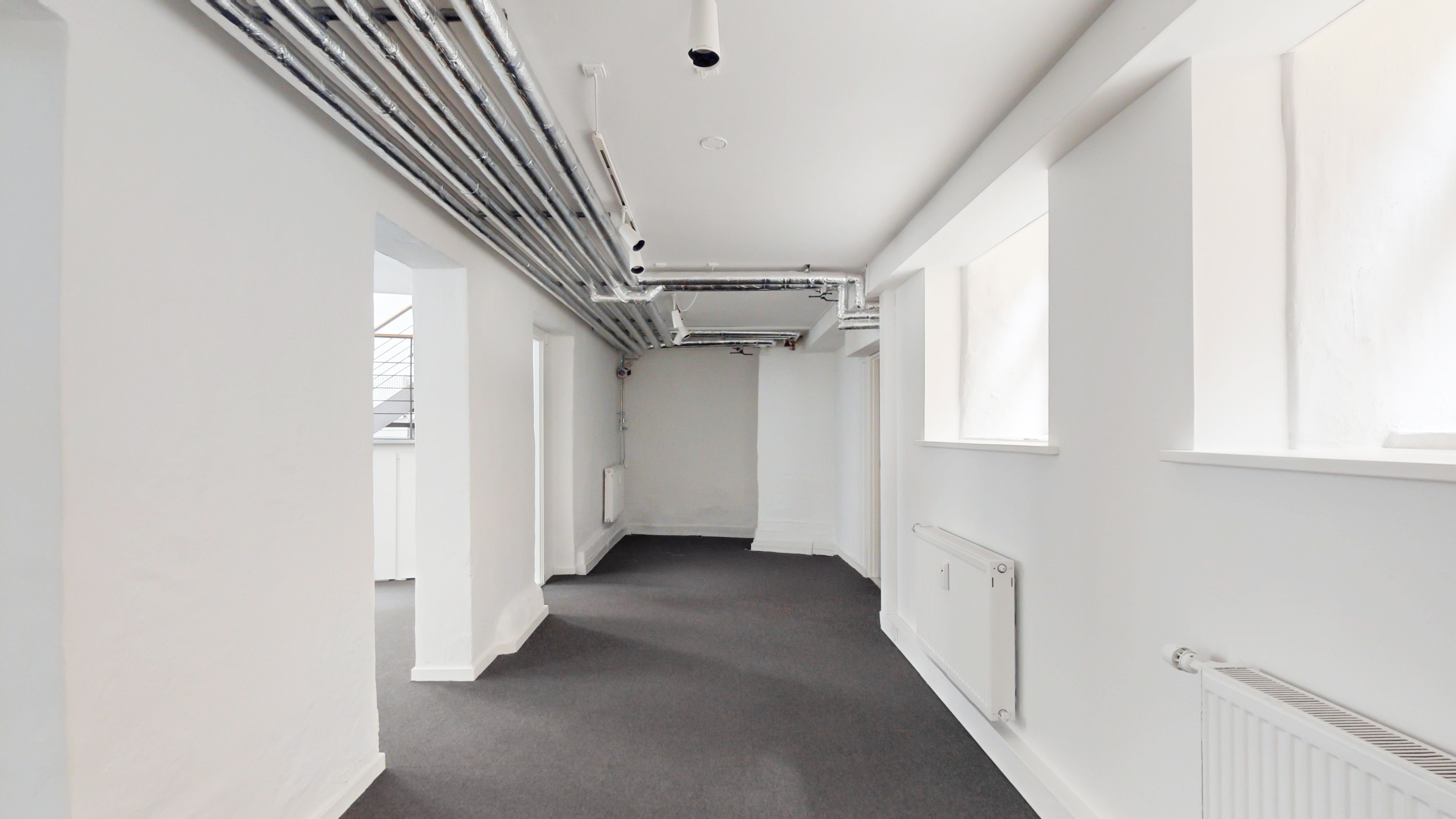 Hvidt lokale med gulvbelægning og hvide vægge, der vender mod hverandre. De to sider er adskilt af hvide søjler med hvide radiatorer. Loftet er hvidt med rør og flere lysekroner.