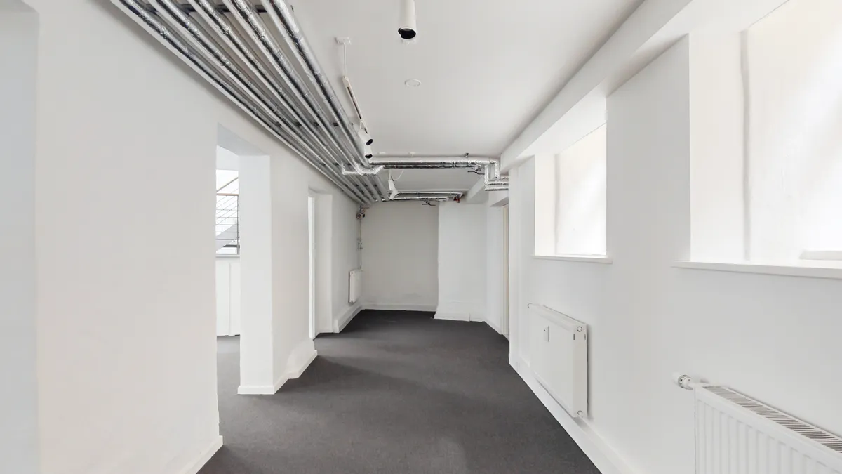 Hvidt lokale med gulvbelægning og hvide vægge, der vender mod hverandre. De to sider er adskilt af hvide søjler med hvide radiatorer. Loftet er hvidt med rør og flere lysekroner.