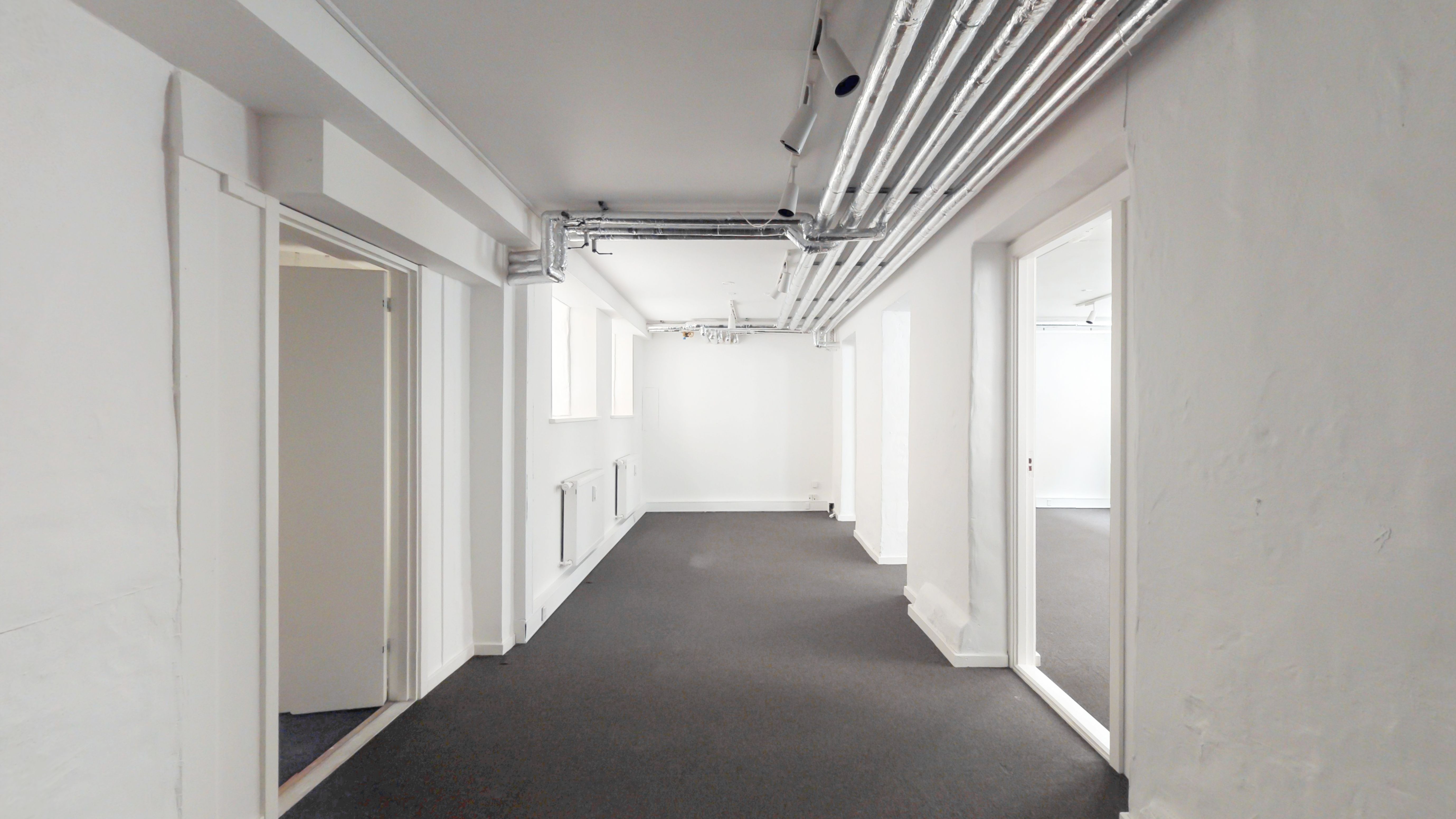 En lang, ubeboet korridor med hvide vægge, grå gulvbelægning og en række hængende rør i loftet