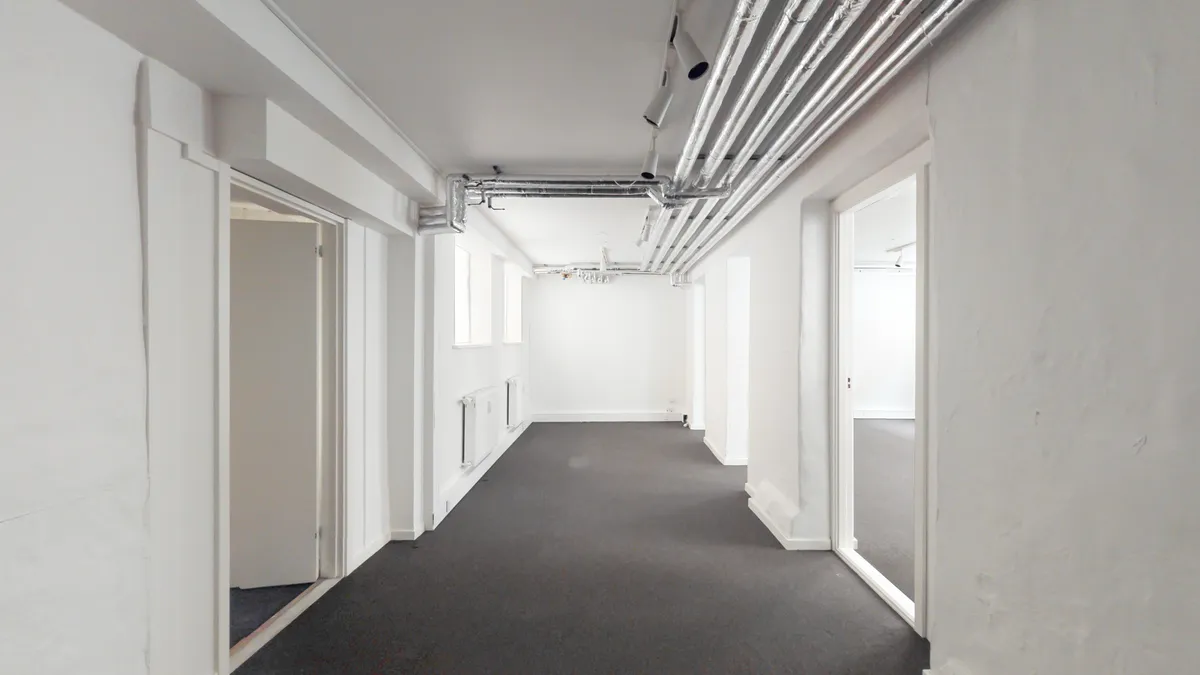 En lang, ubeboet korridor med hvide vægge, grå gulvbelægning og en række hængende rør i loftet