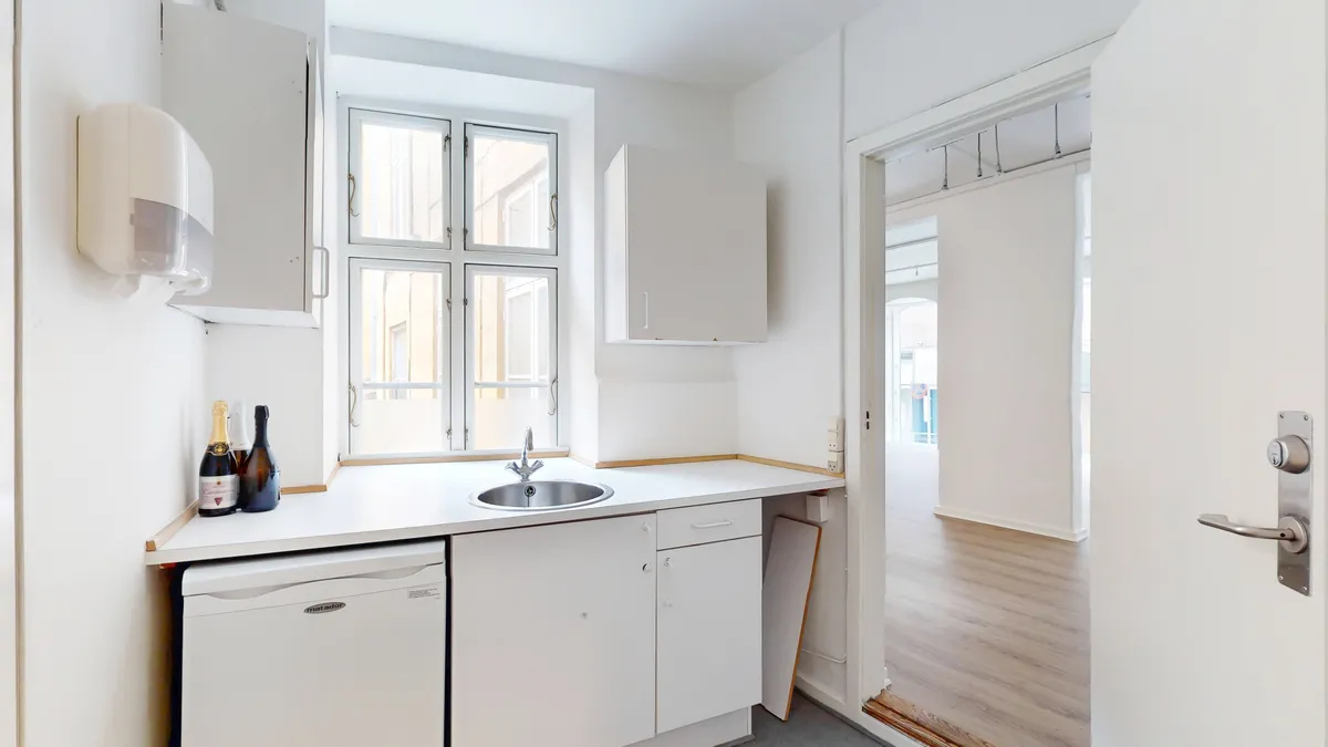 Foto af en minimalistisk køkken i et detaillejemål med køkkenvægge i hvidt og et vindue med solskærm. To champagneflasker står på køkkenbordet. Der er en håndvask med vandhane, og en håndklædeautomat er monteret på væggen.