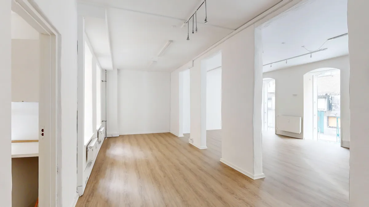 Store, tomme rum med hvide vægge og lysegule gulve. Højloftet med hængende lamper. Store vinduer med lys ind. To hvide radiatorer og åbne døre til mindre rum