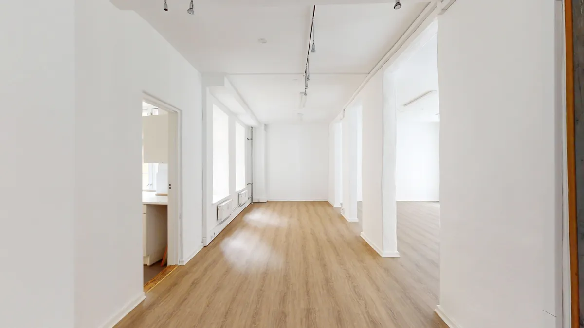 Foto af en stor, tom og lys hall med hvide vægge og gulv. Loftet er hvidt med flere nedhængte lysekroner. Hallen er ubeboet med åbne døre til flere lokaler.