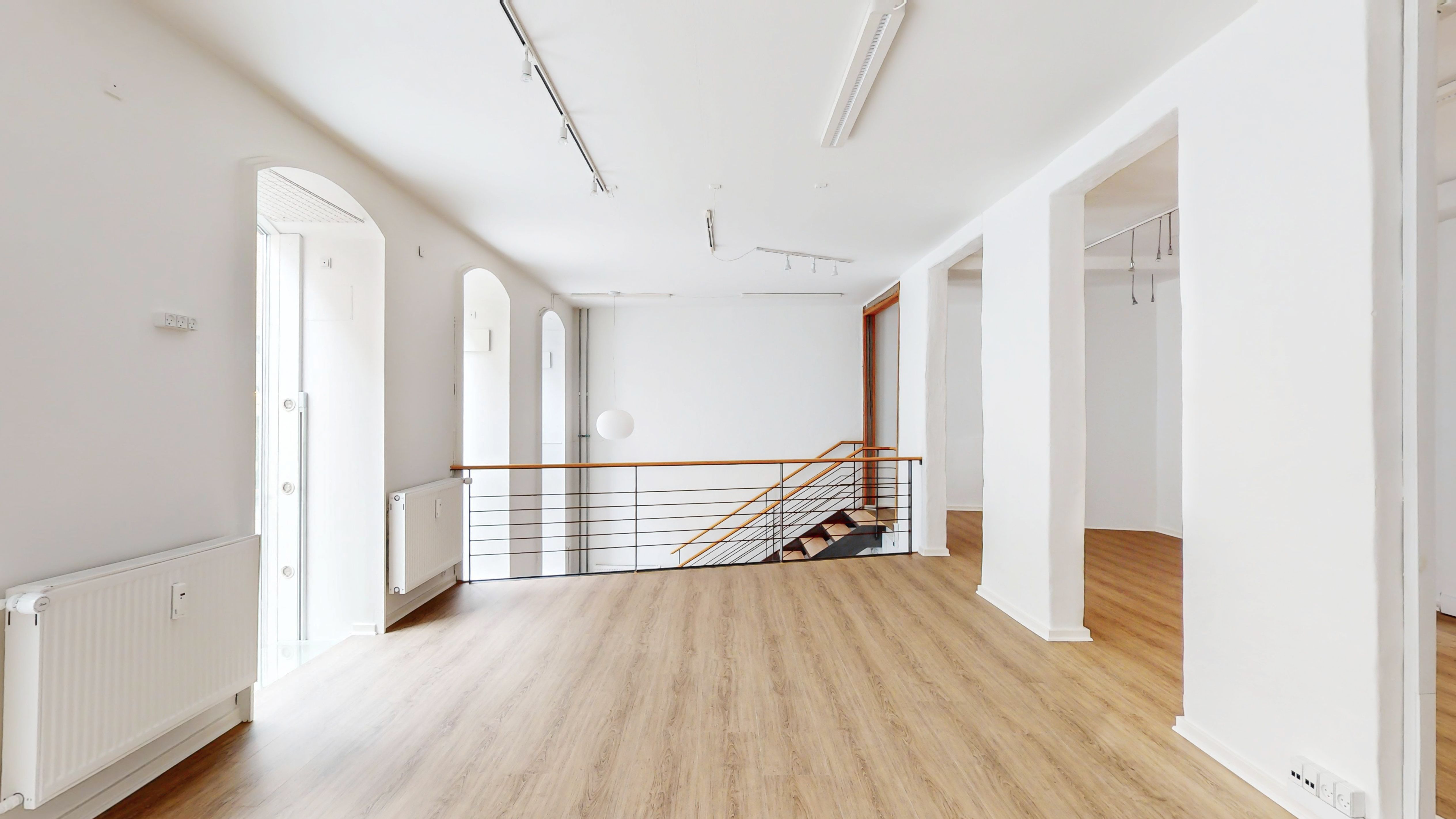 Forretningslokal med lys og rummeligt mødelokale. Indeholder slanke hvide vægge, lysegulve og moderne radiatorer. Loftet er højt og lysekroner hænger elegant. Der er et slankt trappeværk med metalgevær.