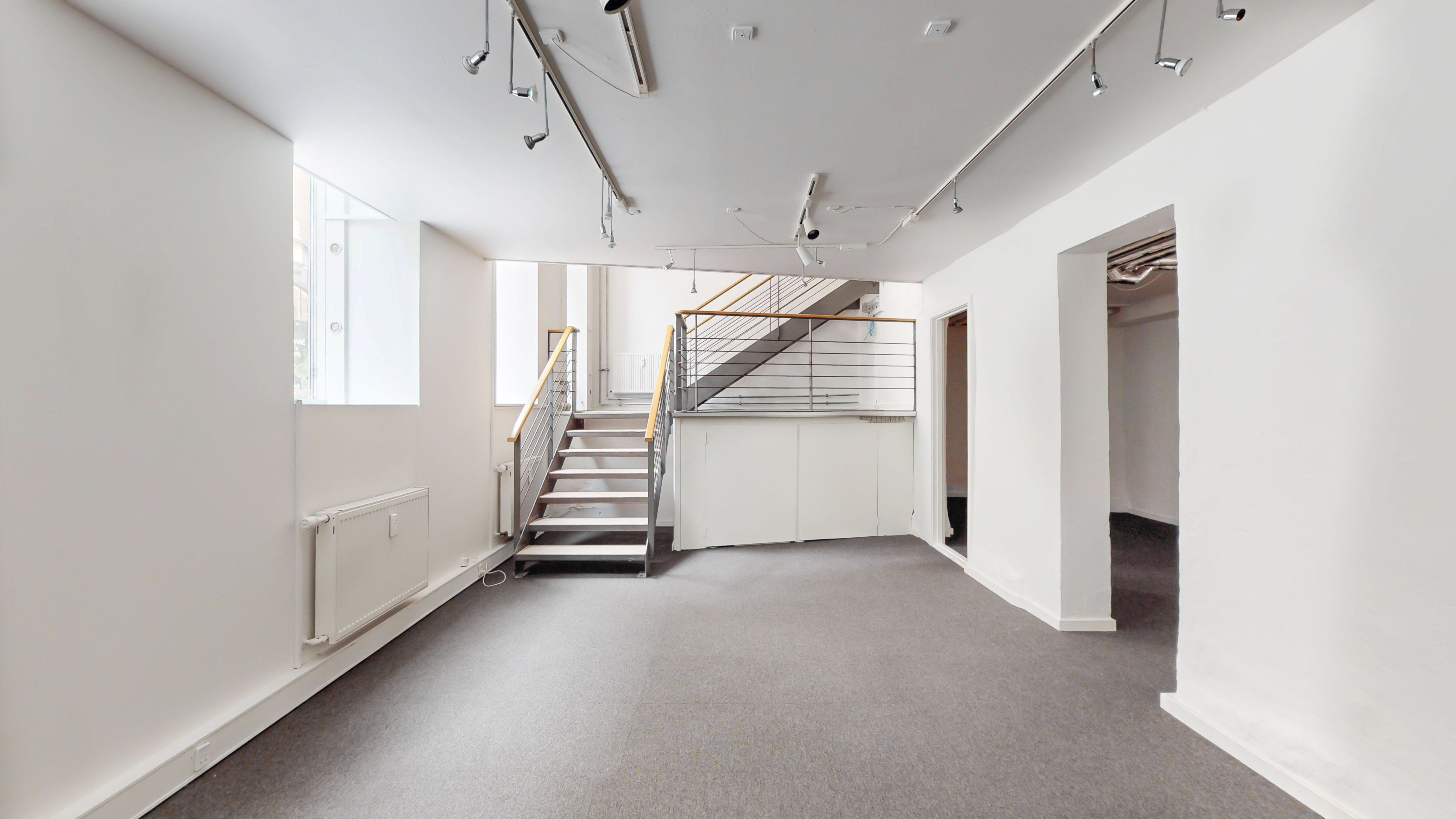 Butikslokale i stueplan med hvide vægge, gulv og loft samt et stigeanlæg med metalgesims og gult håndtag.