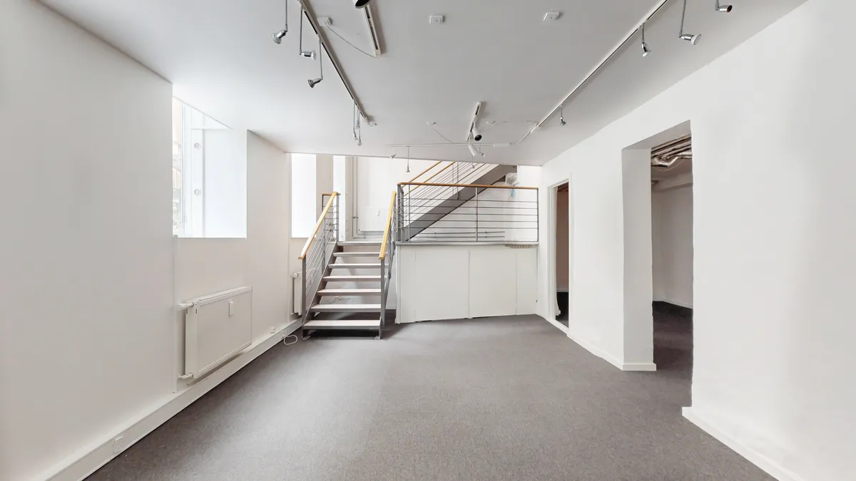 Butikslokale i stueplan med hvide vægge, gulv og loft samt et stigeanlæg med metalgesims og gult håndtag.