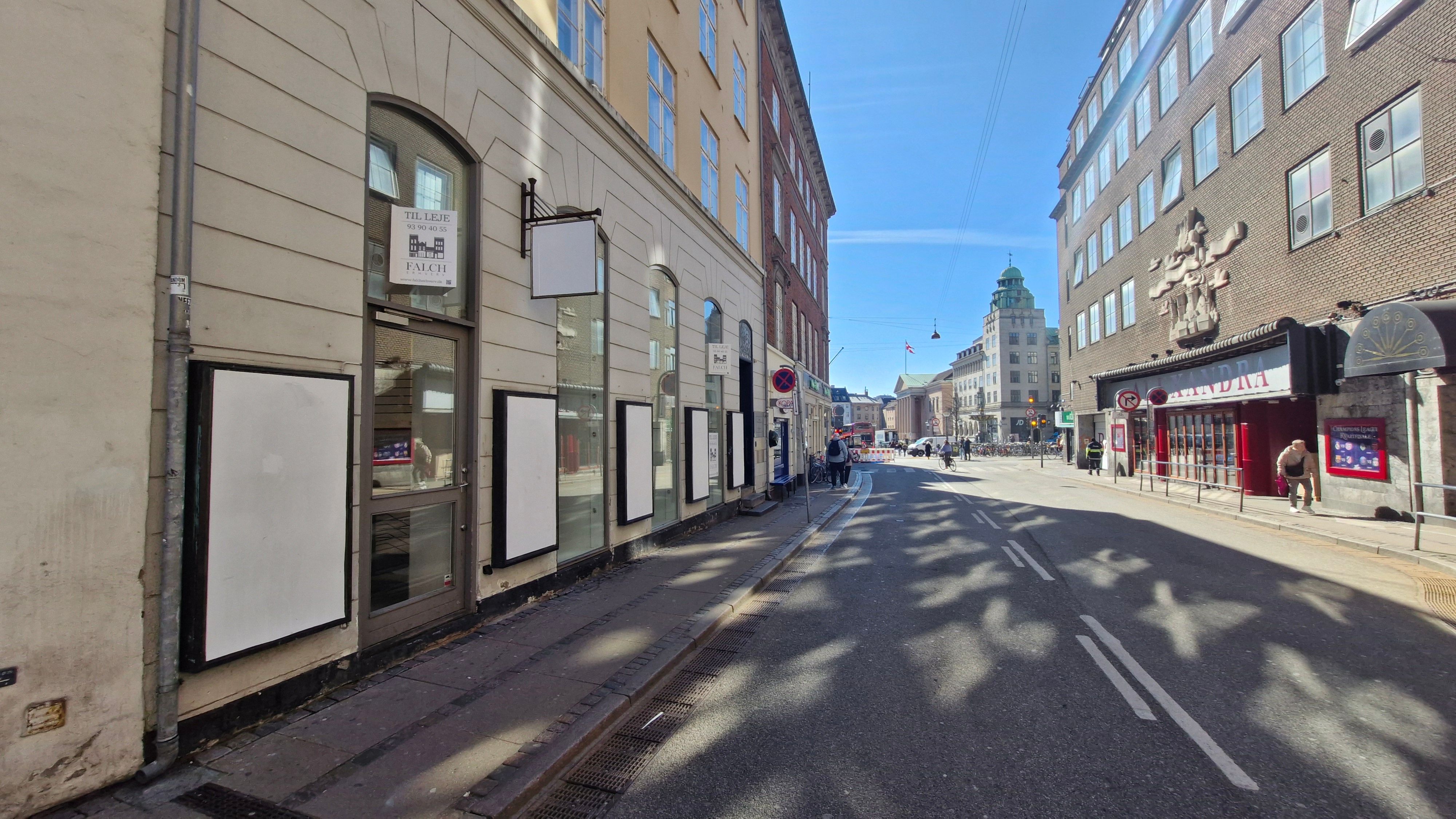 En tom gade i Indre By med tomt butikslokale med skiltet Til Leje. På hjørnet er Københavns Rådhus med rødt flag. En person går på fortovet og bærer en rygsæk