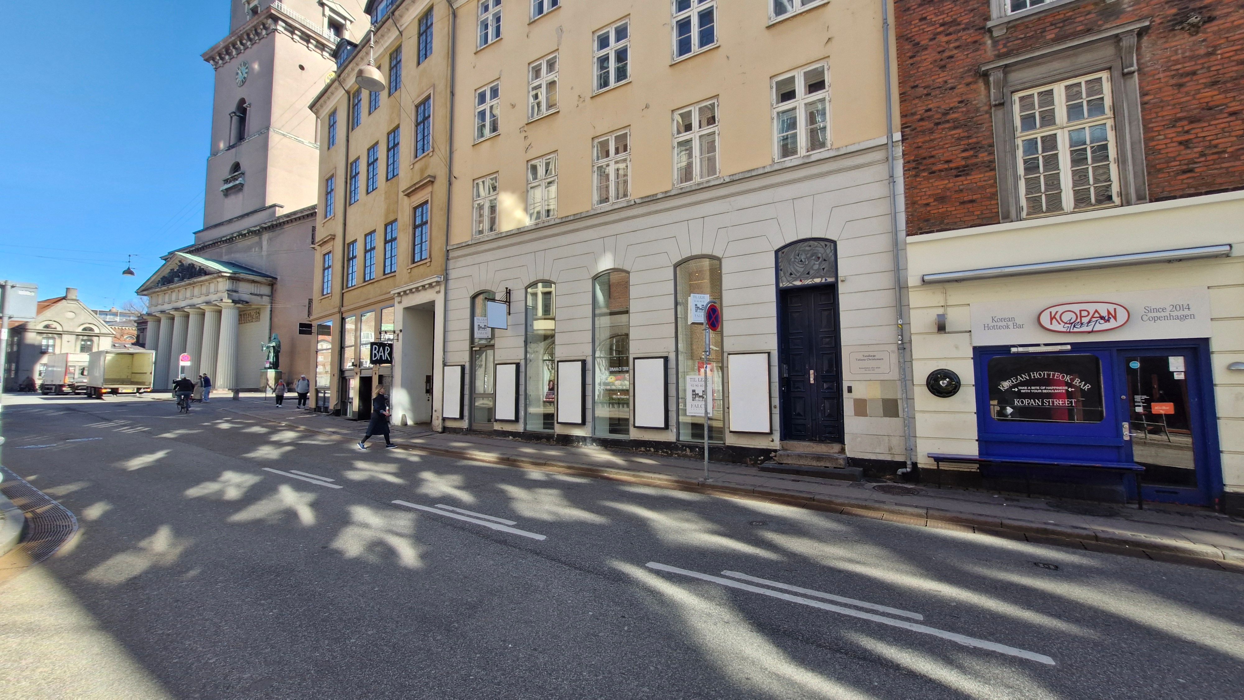 På Nørregade 4 i København K er der et stort hvidt og gult lejemål med flere indgange og vinduer. Lejligheden vender ud mod en tom gade med mange skygger og en kvinde på cykel