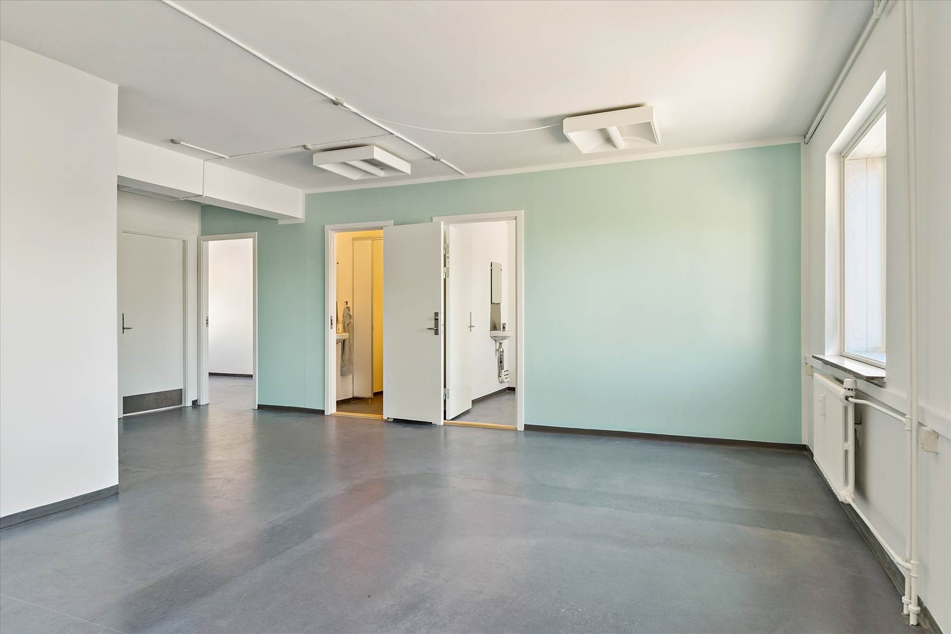 En tom, lyseblå stue med hvide vægge, lysegrå gulv og åbne døre til andre værelser og en radiator