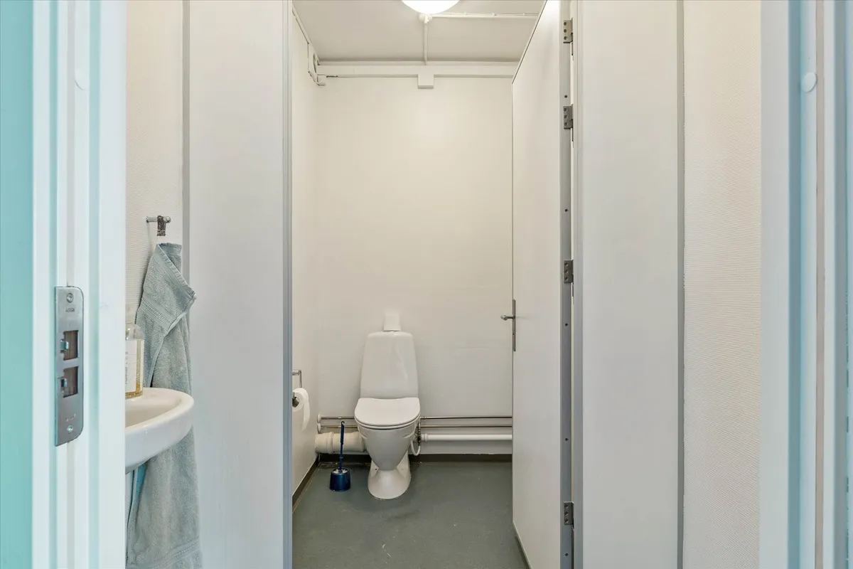 En toiletrum med toilet, sækkeholder og håndklæde. Toiletrummet er smal og lille med hvide vægge og gulv samt et hvidt