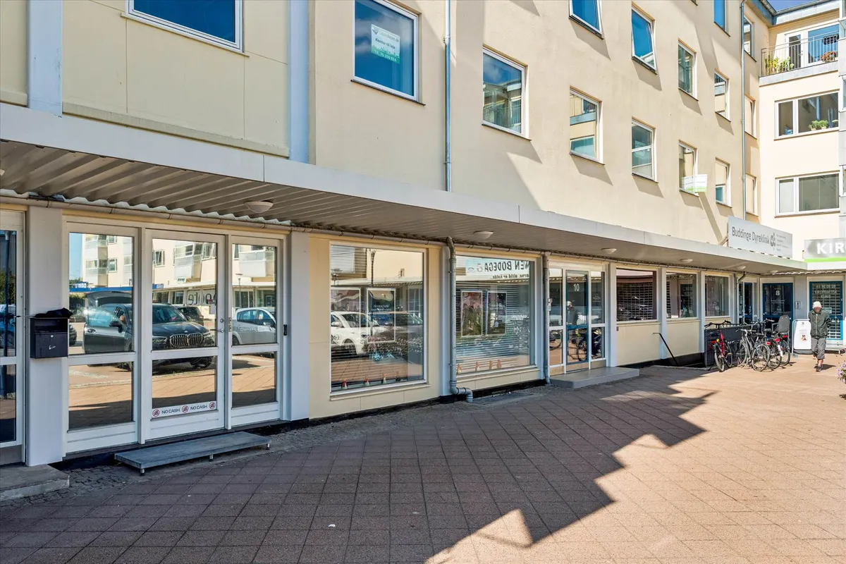 Lejemål på Bagsværd Torv 10 med butikslokaler og bilparkering. Kørecykler og en voksen mand står udenfor.
