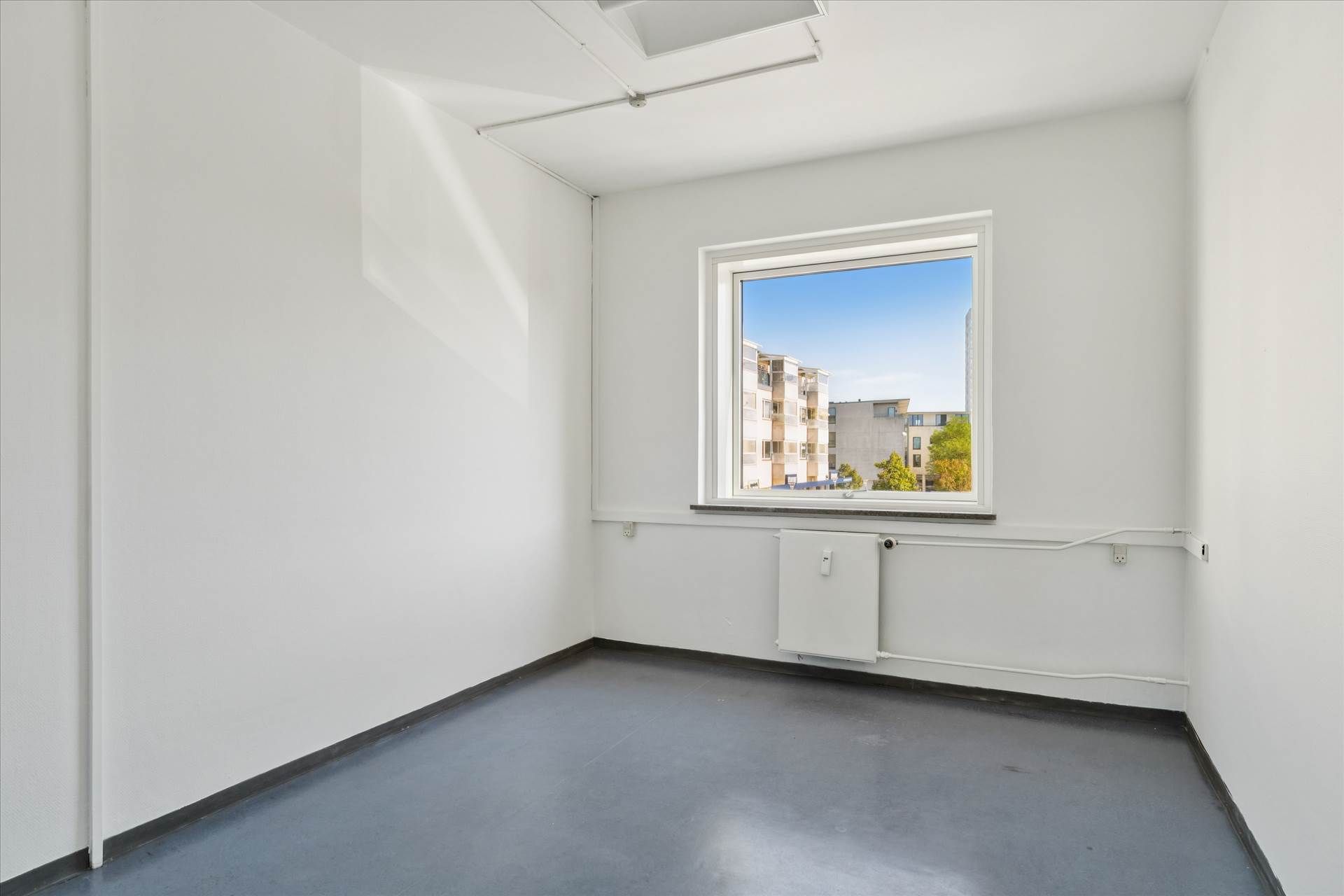 Lukket rum med hvide vægge og gulv og et vindue med udsigt til bygninger og grønne træer