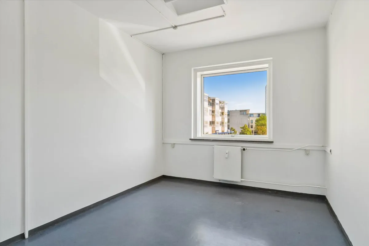 Lukket rum med hvide vægge og gulv og et vindue med udsigt til bygninger og grønne træer