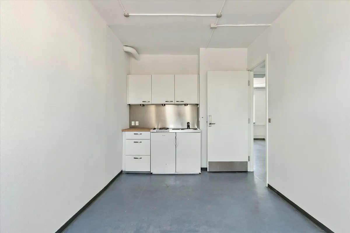 Lys og ren stue med hvide vægge og loft samt gulv, køkken med brædder, strygejern og køleskab samt lysekroner og åbne døre