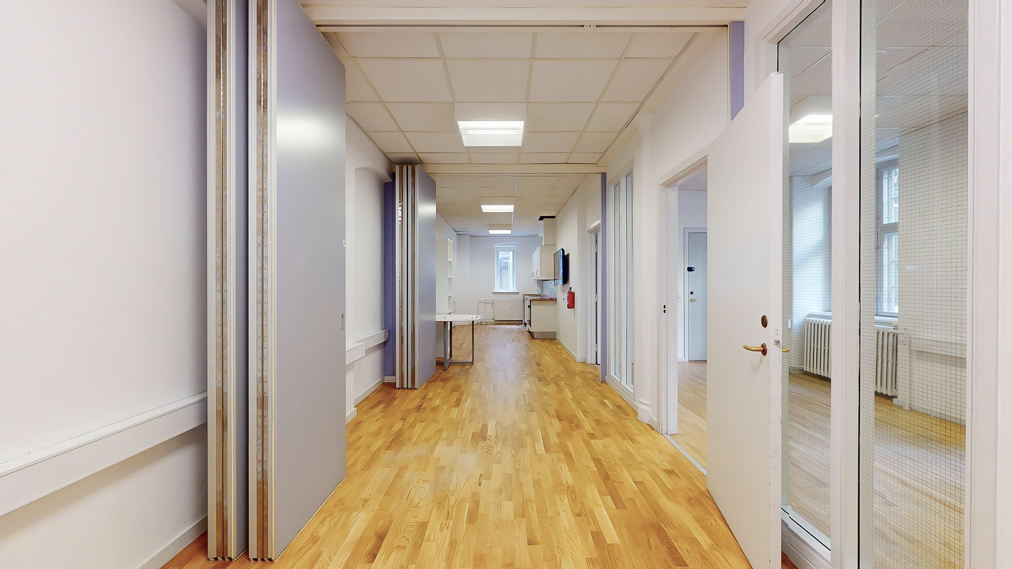 En lyse og moderne korridor med trægulve og hvide vægge og vinduer med lysegardiner og et rødt brandslukkerapparat.