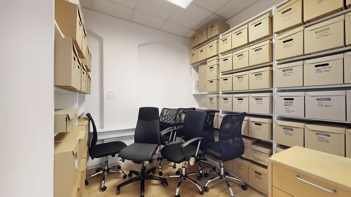 Kontor med høje hylder og mange kartoner med mærkning, der viser datoer og indhold. Der er fire ergonomiske kontorstole med