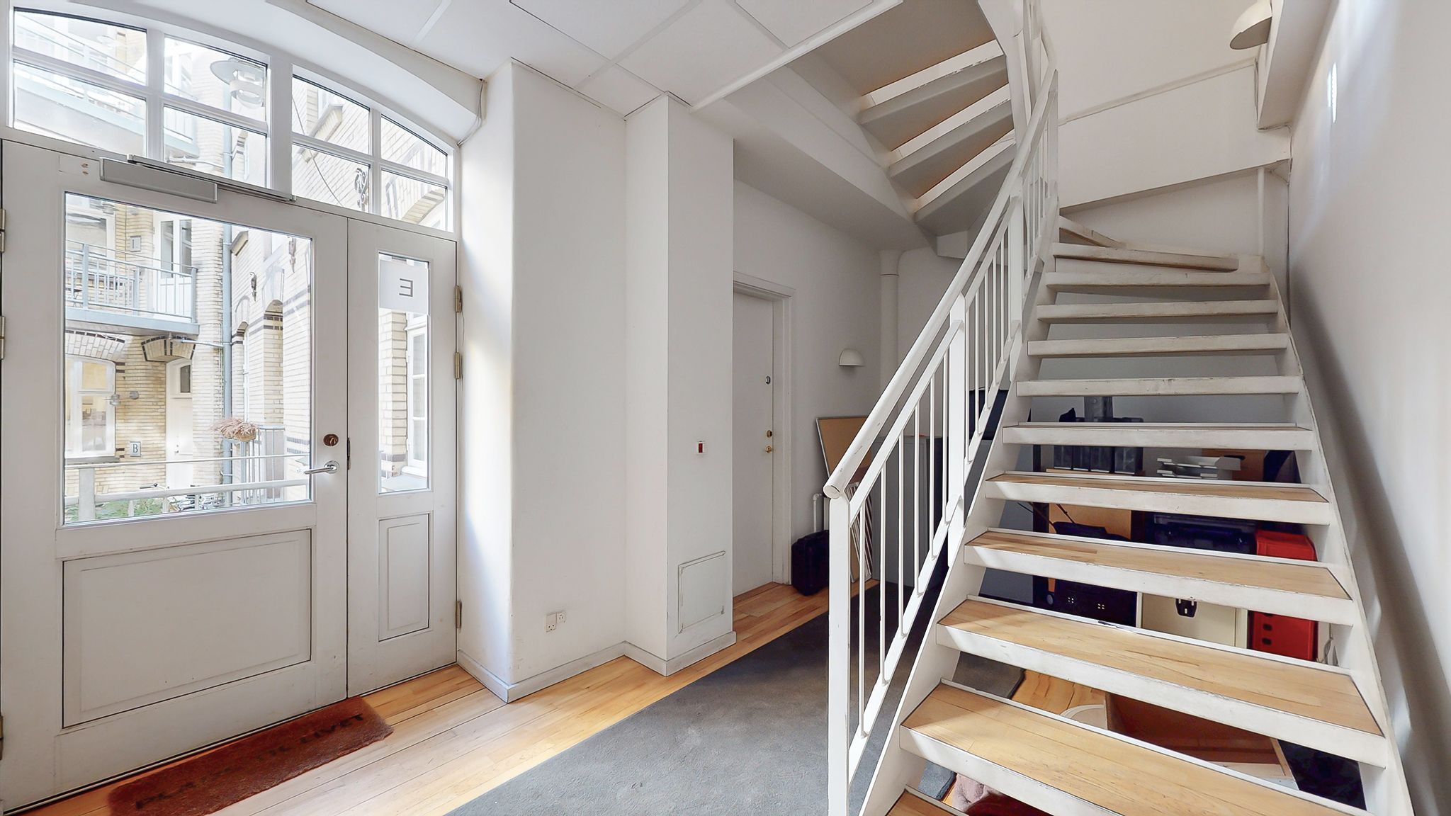 Indtræd i en velrenoveret og lyse stue med hvide vægge og trægulve samt lyse trappetrin og hvidt håndtag