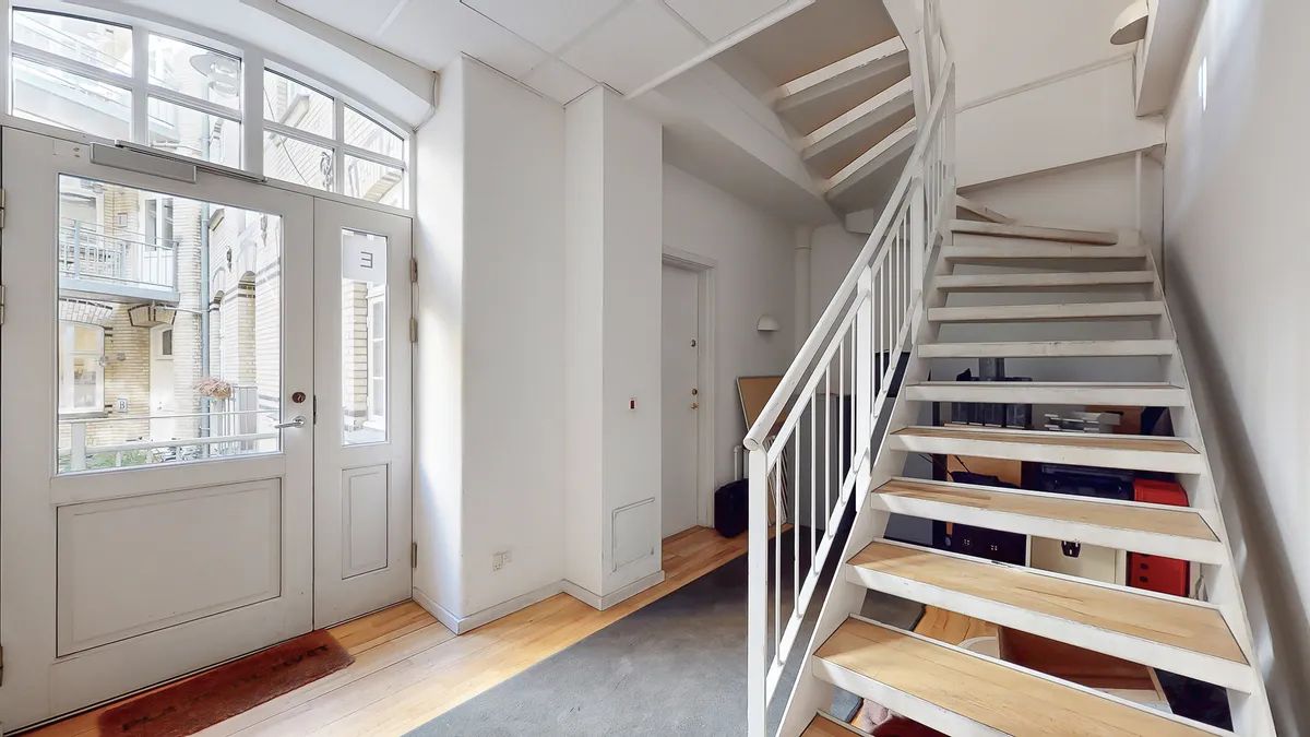 Indtræd i en velrenoveret og lyse stue med hvide vægge og trægulve samt lyse trappetrin og hvidt håndtag