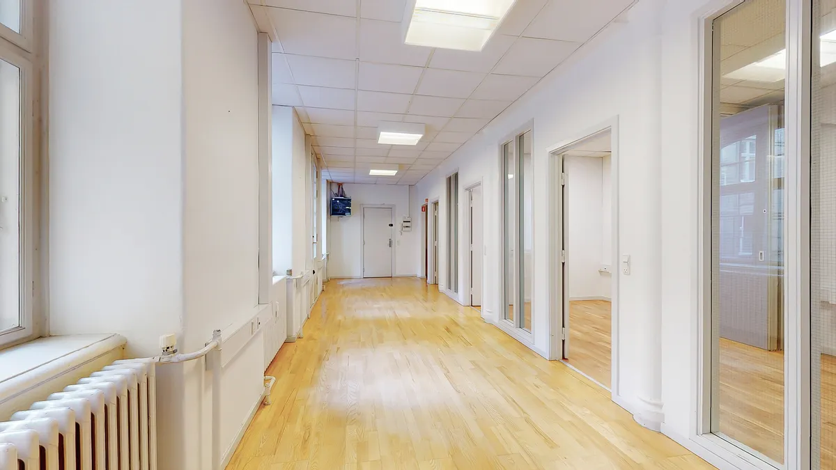 Et langt og smalt kontorlokale med hvide vægge og lysegrå loftsplader. Gulvet er lavet af lyse træplanker. Der er radiatorer