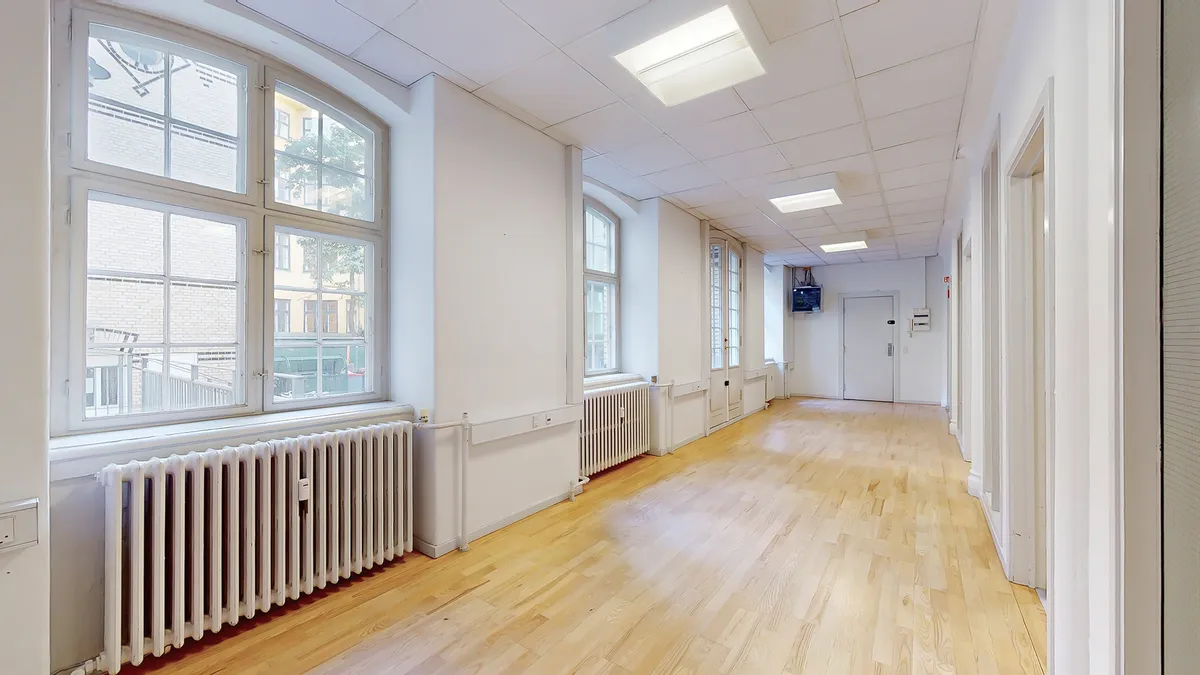 Kort og lyst kontor med hvide vægge og lyse gulve samt store vinduer med udsigt til bygninger i baggrunden