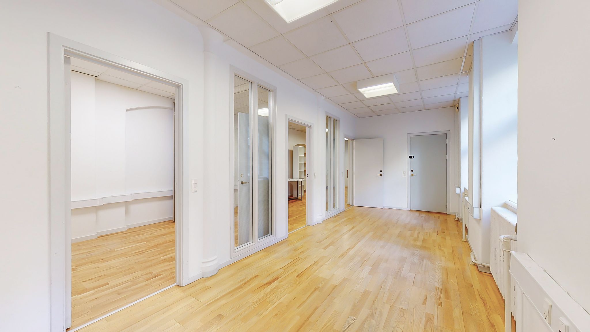 En stor og lys hall med hvide vægge og lysegrå gulv. En del af det lyse gulv er dækket af en møbel. Hallen har flere døre og