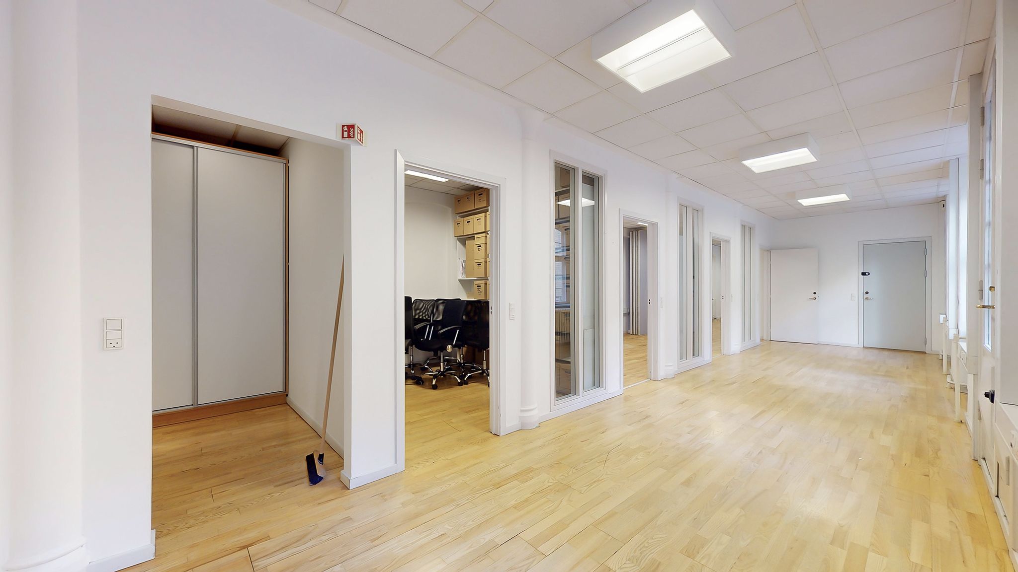 Kontor med lyse gulvbelægning og hvide vægge, hvor der er flere glasdøre og åbne døre til en mindre rummelig gang.