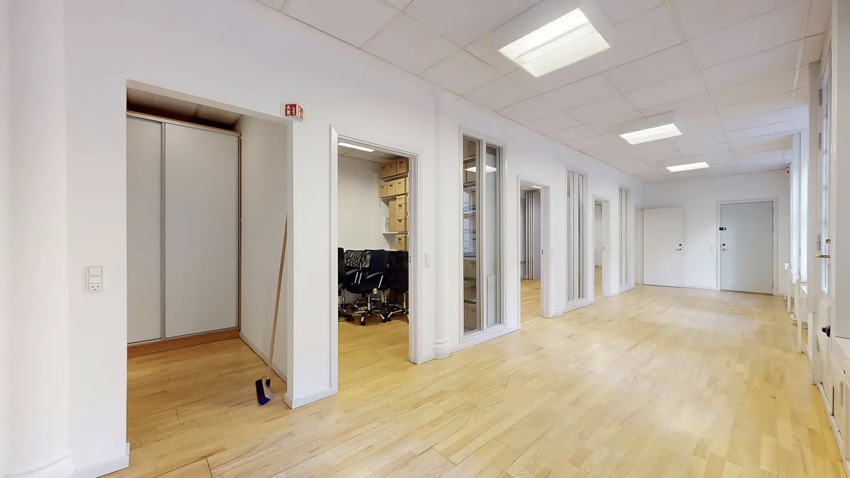 Kontor med lyse gulvbelægning og hvide vægge, hvor der er flere glasdøre og åbne døre til en mindre rummelig gang.